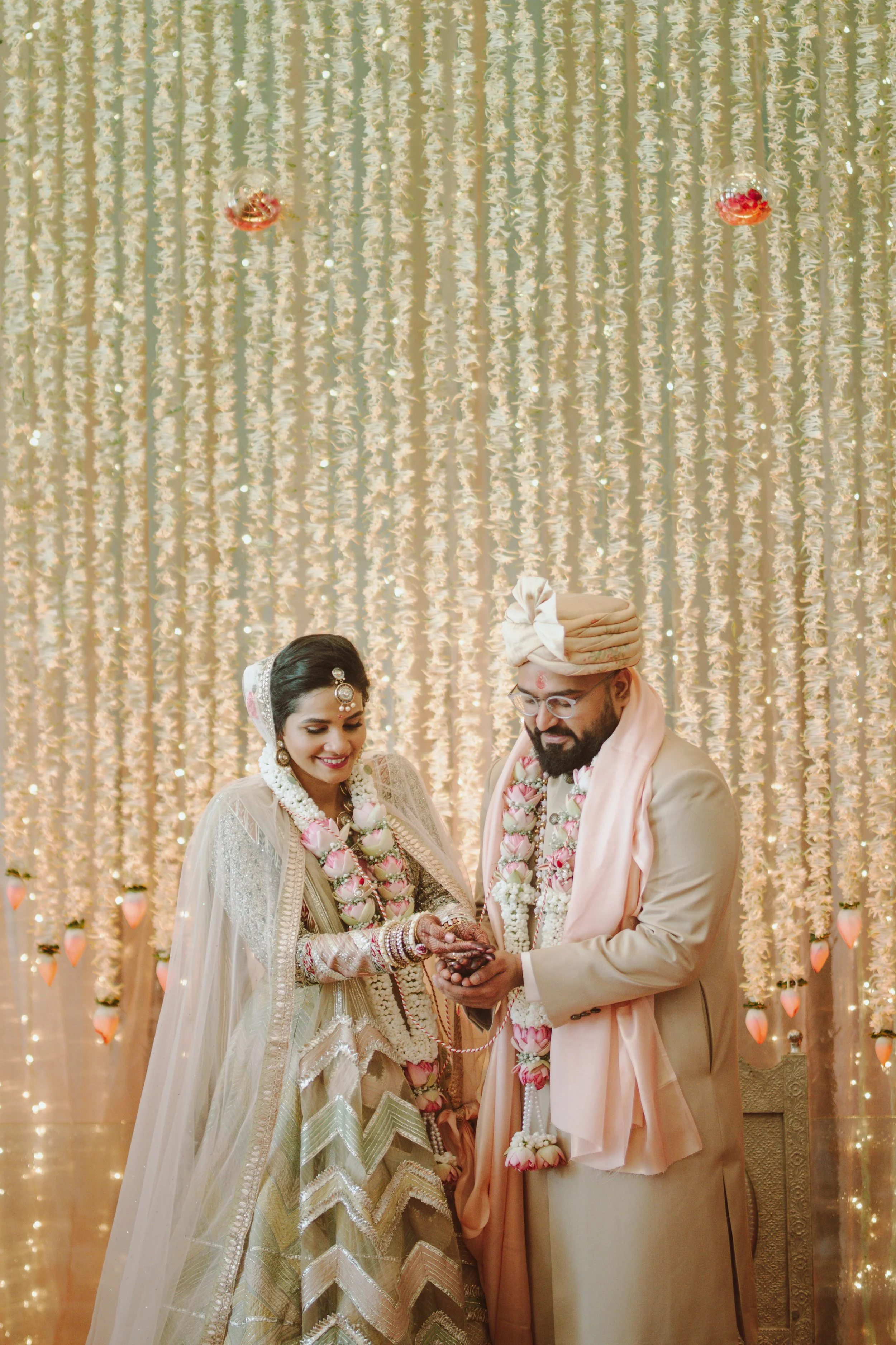 A bride and groom in traditional Indian wedding attire exchange rings in front of a decorated backdrop.