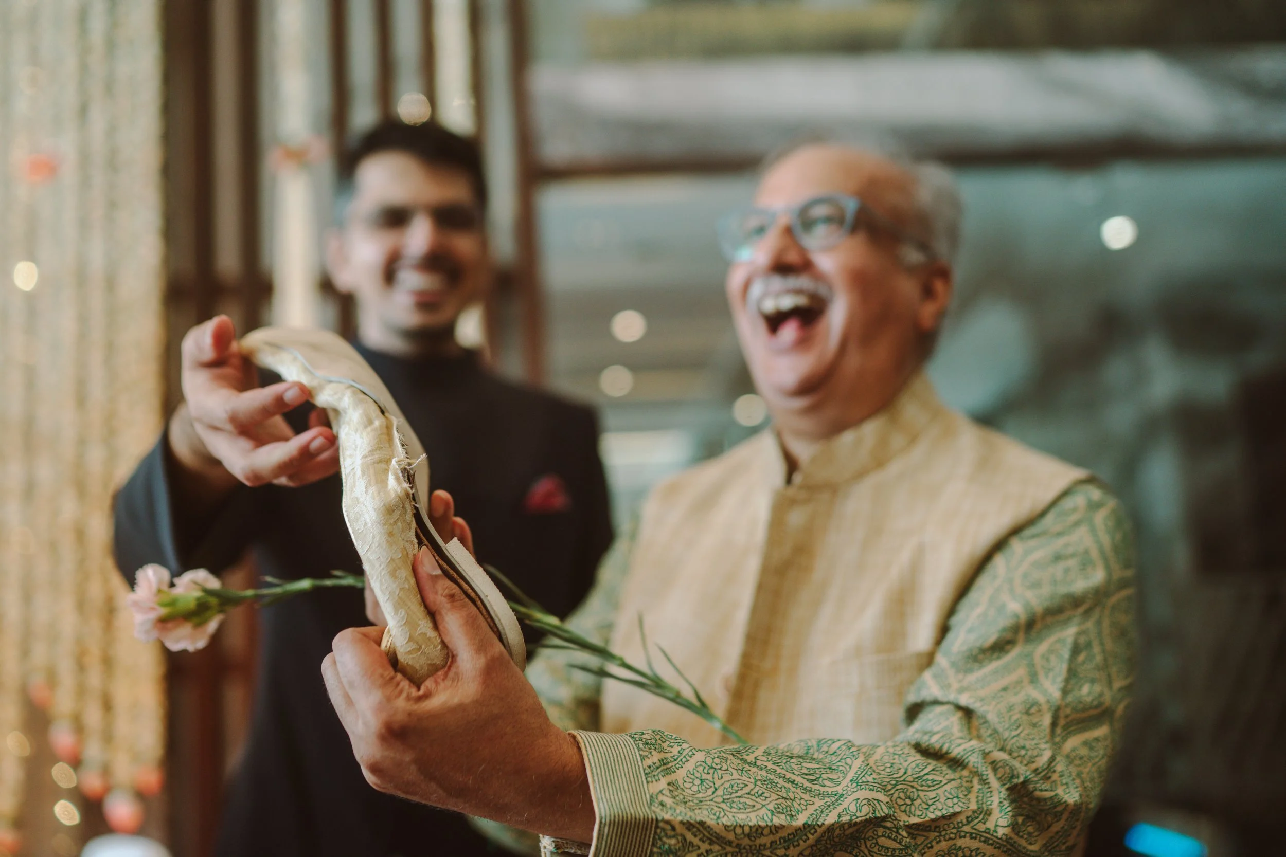 Two men, one older in traditional Indian attire and one younger in a black blazer, laughing and smiling indoors. The older man is holding a snake, which the younger man is touching.