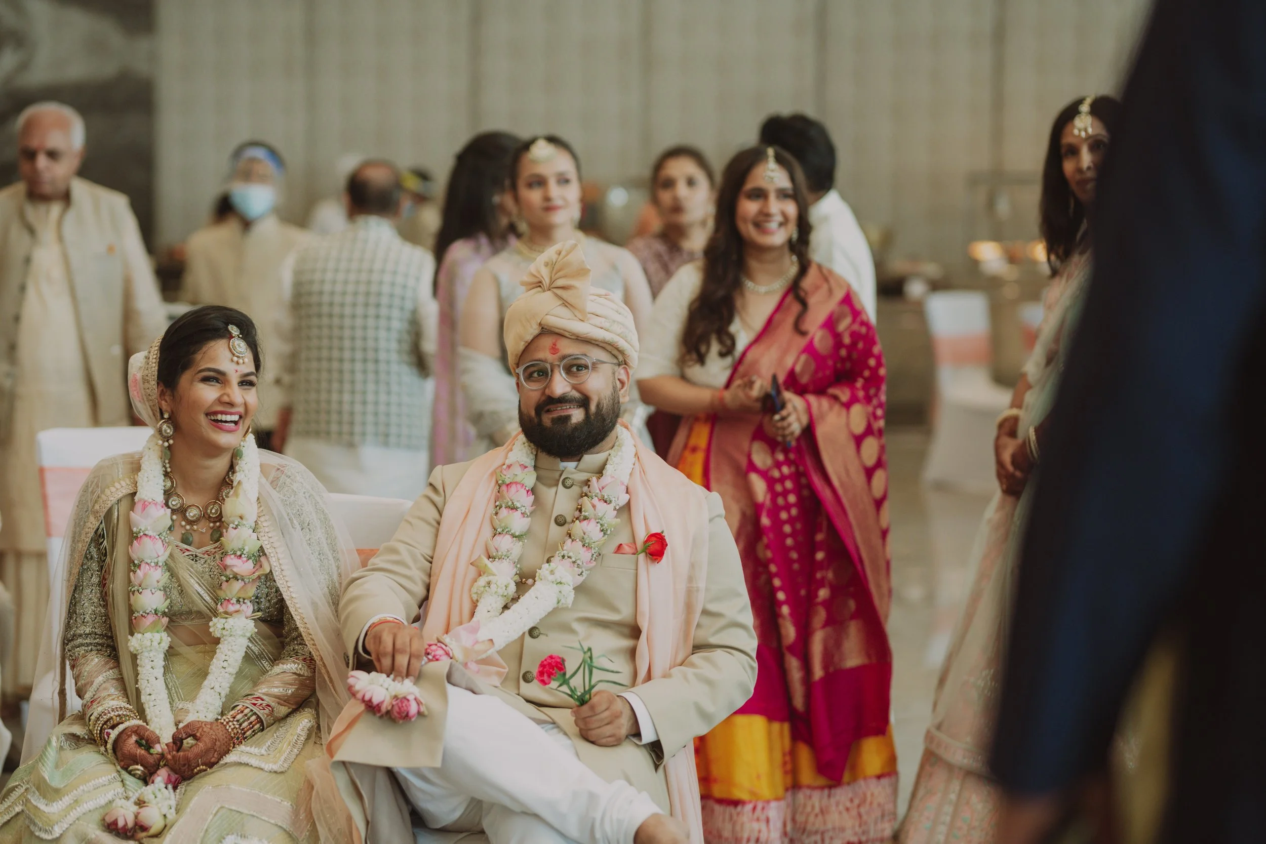 Indian wedding ceremony with the bride and groom seated, smiling, and surrounded by family members dressed in traditional attire.