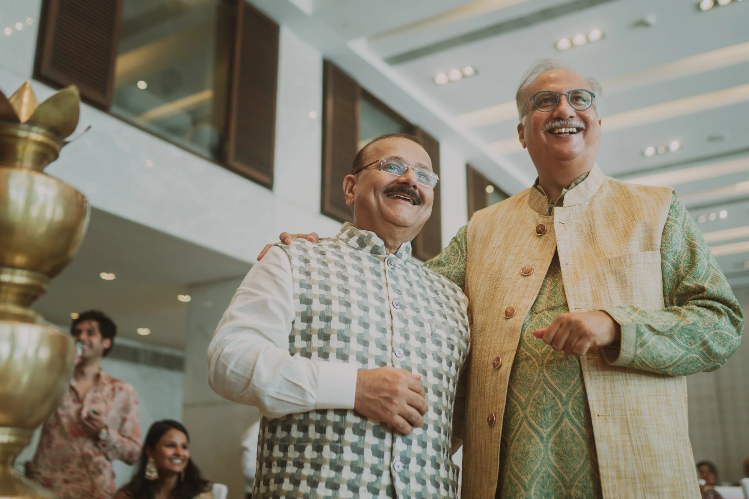 Two men in traditional Indian attire smiling and posing together at a celebration or event, with other people in the background.