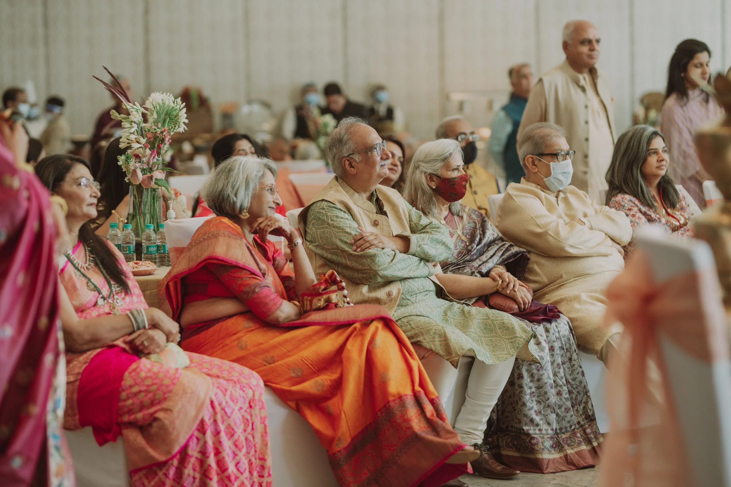 A group of adults seated and watching an event, some wearing traditional Indian clothing and face masks, in a decorated indoor setting.