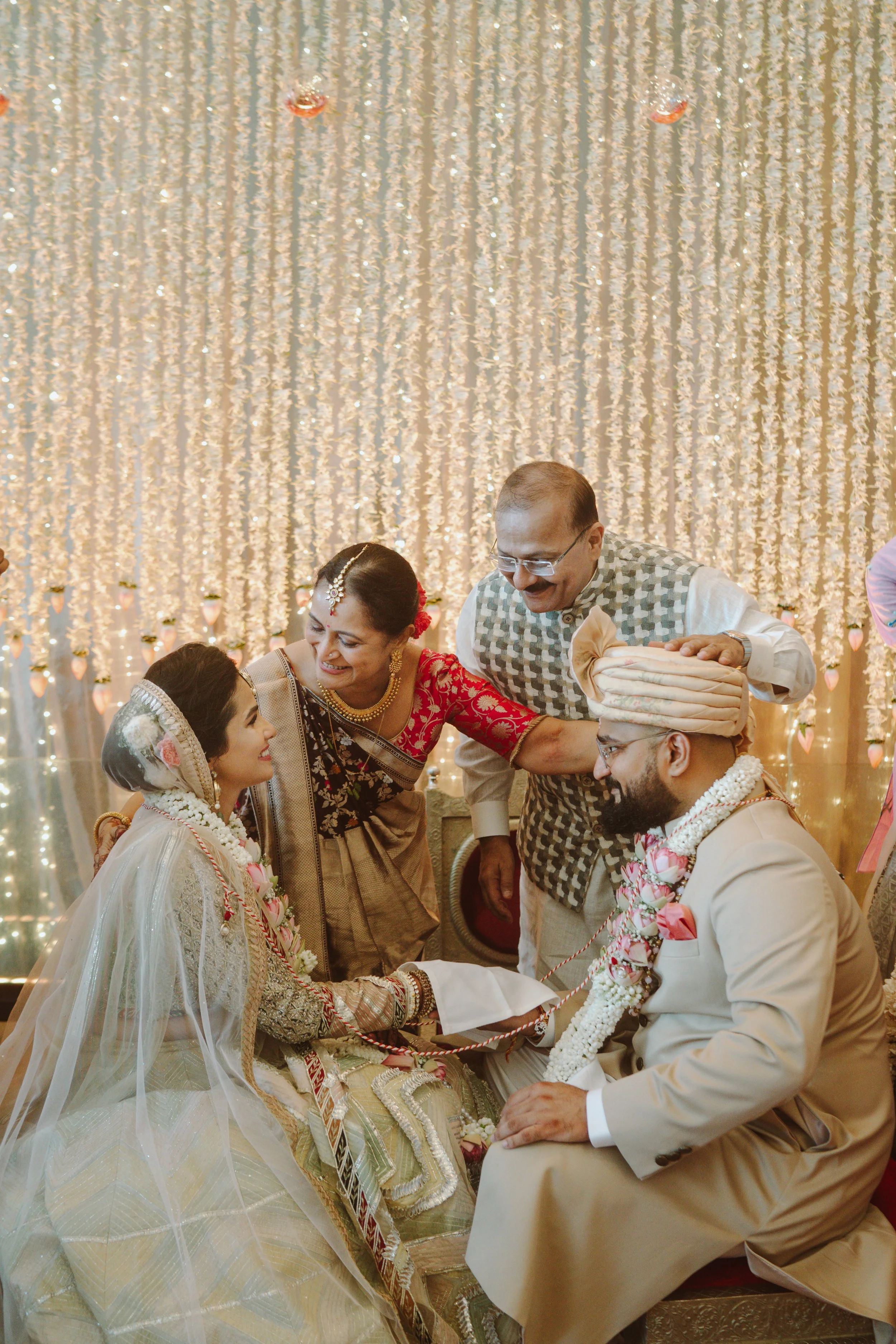 Indian wedding ceremony with bride and groom seated, surrounded by family members, in front of a decorated backdrop of lights and flowers.
