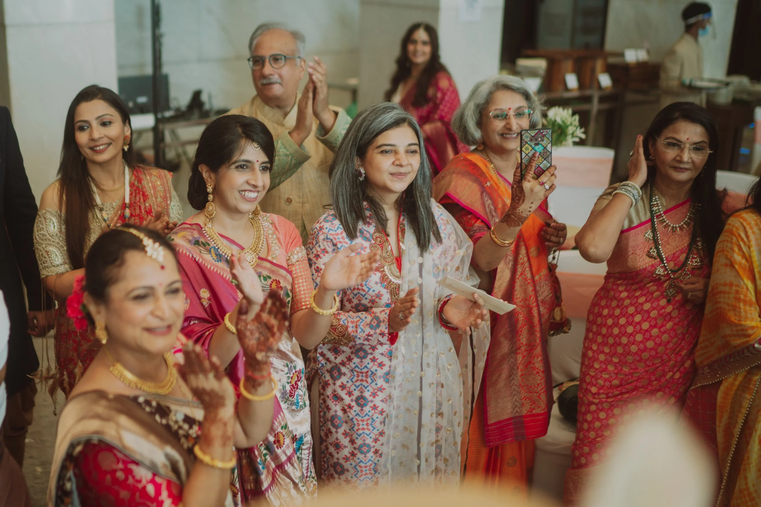 Group of women and a man in traditional Indian attire at a celebration, some clapping, smiling, and one woman taking a photo with a smartphone. In the background, there is a woman in a red outfit and a hospital staff member wearing a face mask.