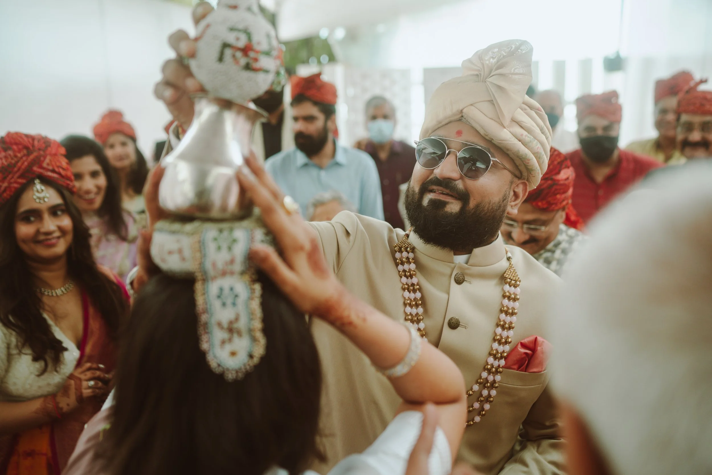Man with a turban, sunglasses, and traditional Indian attire holding a decorated pottery item during a celebration surrounded by smiling people in colorful traditional Indian clothing.