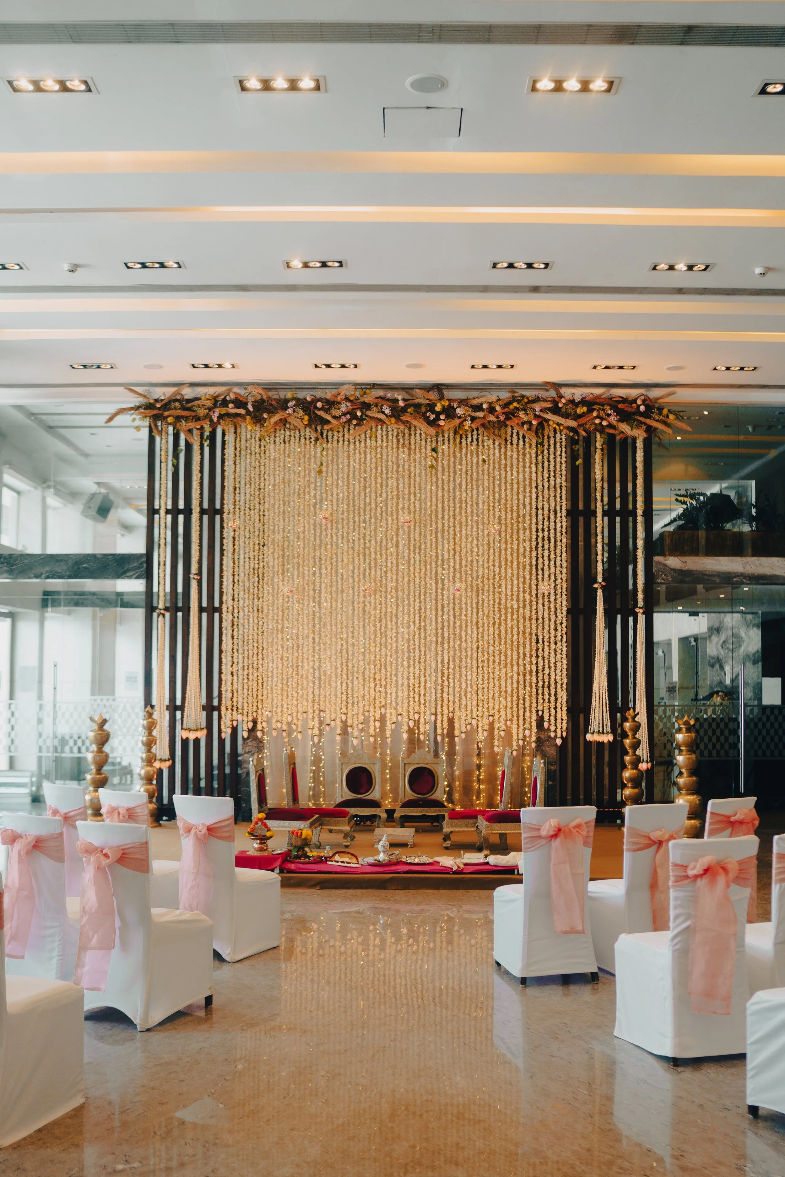 Decorative wedding stage with a gold curtain backdrop, floral arrangements, traditional chairs, and white chairs with pink bows in a modern banquet hall.