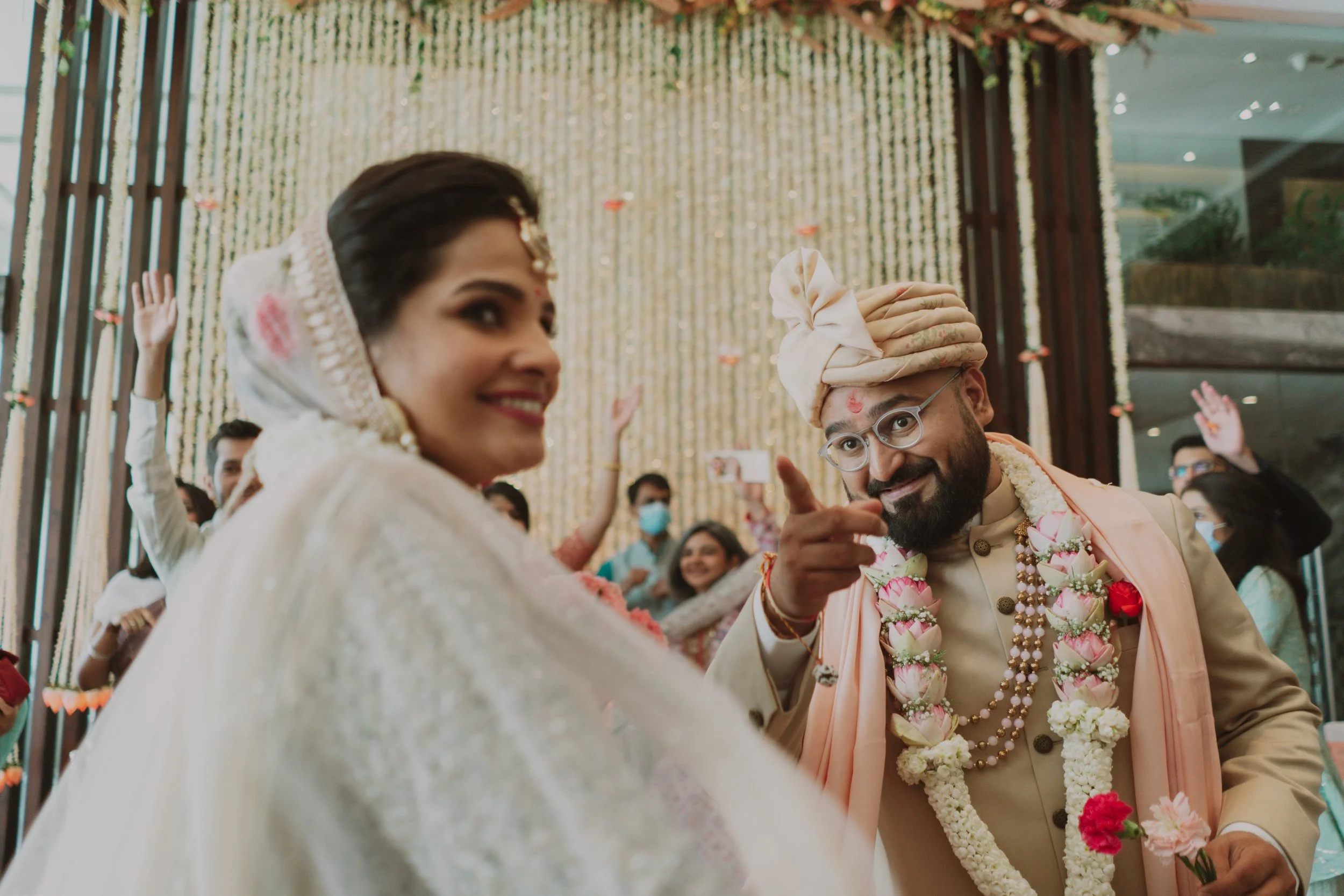 A cheerful bride and groom in traditional Indian attire at their wedding celebration surrounded by friends and family, with a festive decorated backdrop.