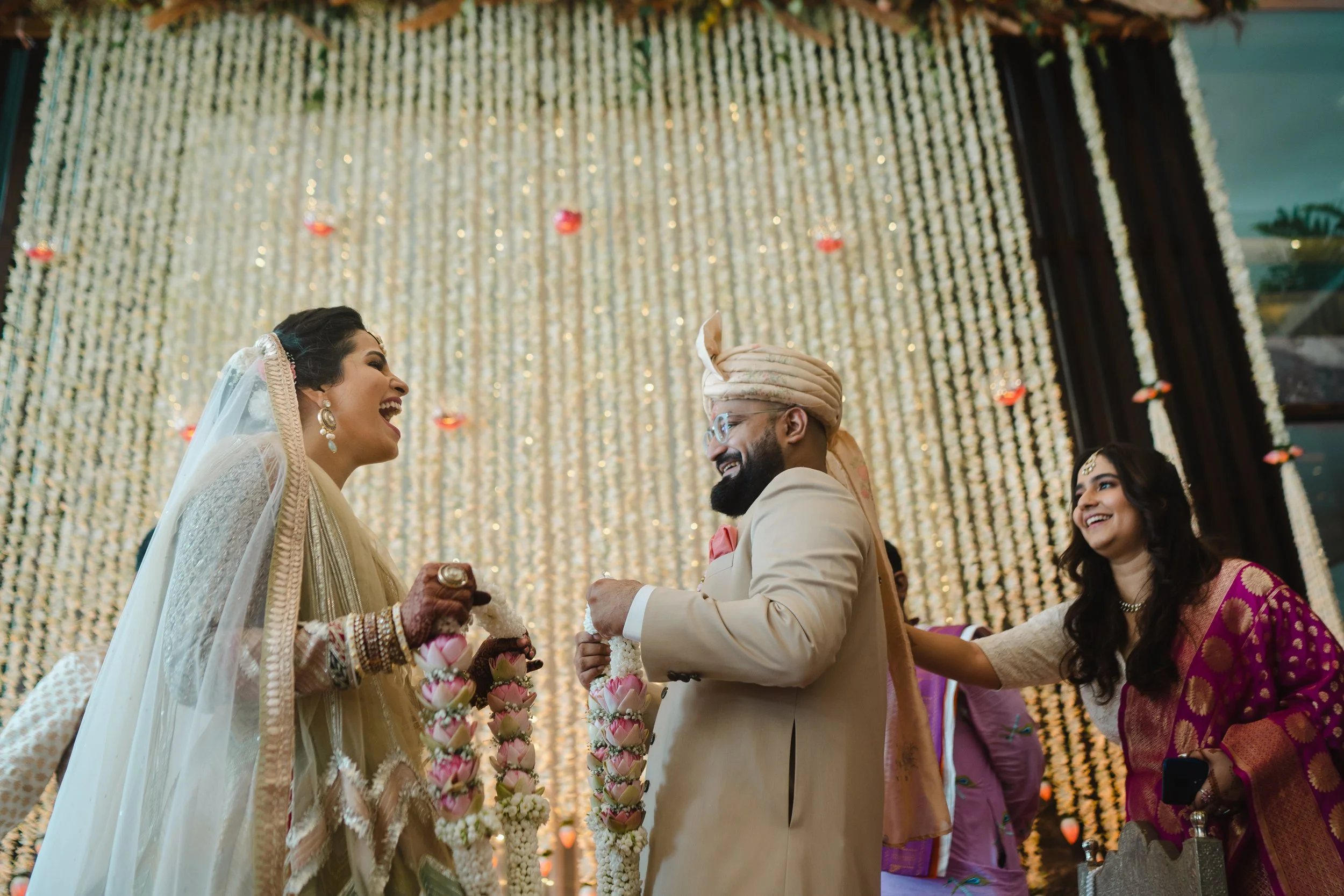 A bride and groom sharing a joyful moment at their wedding, with a woman in a pink and gold sari smiling nearby. The backdrop features hanging white flowers and small floating candles.