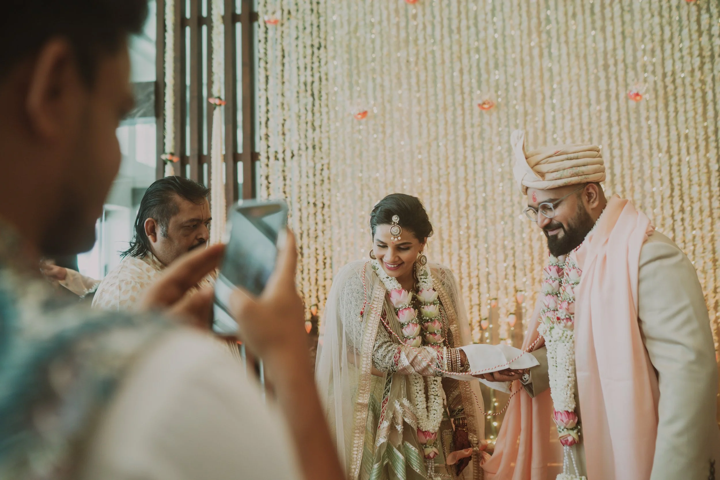 Indian bride and groom at their wedding ceremony, surrounded by family, with a decorated backdrop of hanging flower garlands and warm lights.