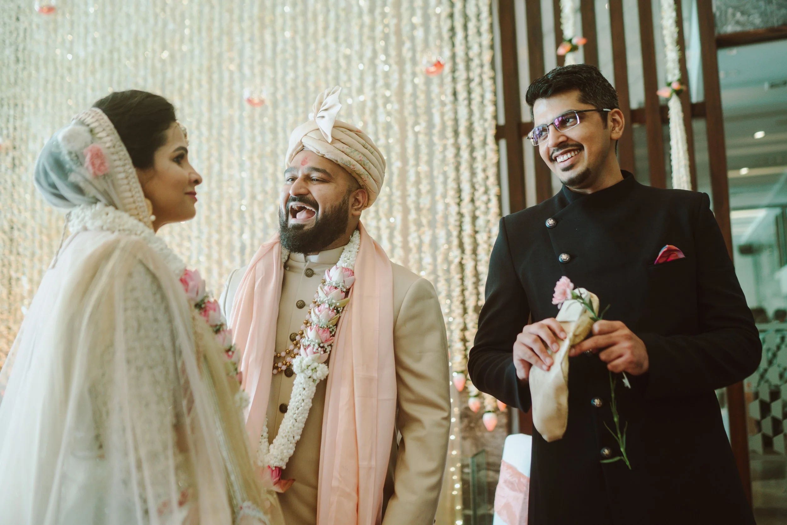 Indian wedding ceremony with bride and groom facing each other, priest smiling, and another man holding a rose.