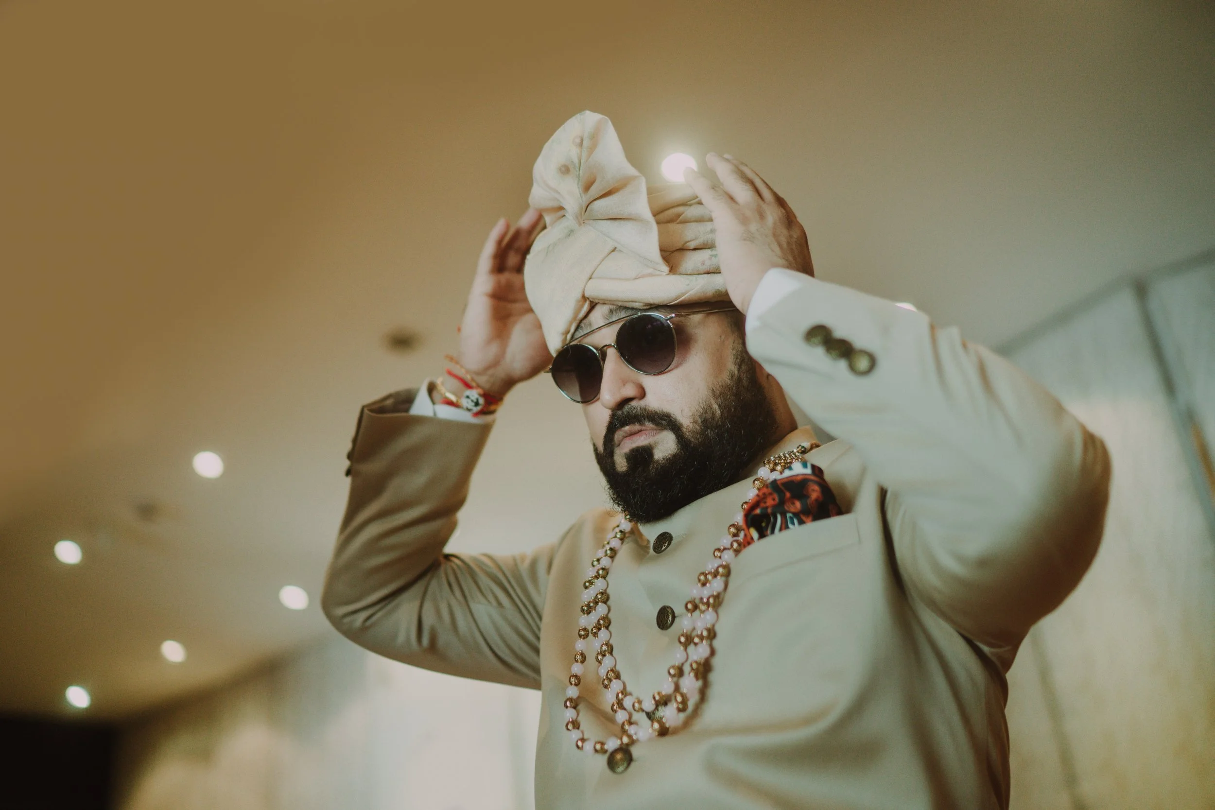 A man dressed in traditional Indian wedding attire, wearing sunglasses and a ceremonial turban, adjusting it with both hands.