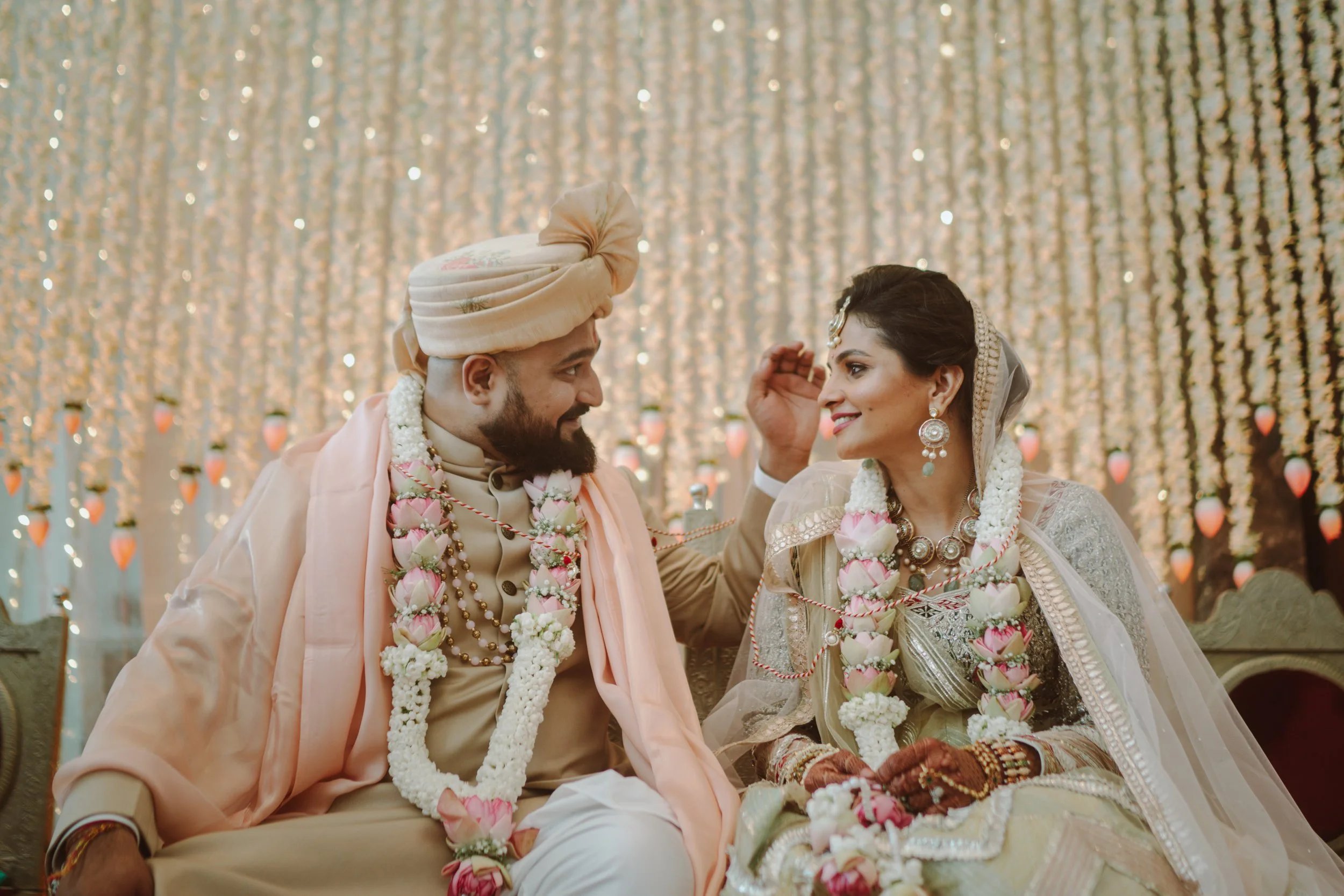 A bride and groom in traditional Indian wedding attire sitting together, smiling at each other, against a backdrop of hanging lights and candles, adorned with floral garlands.