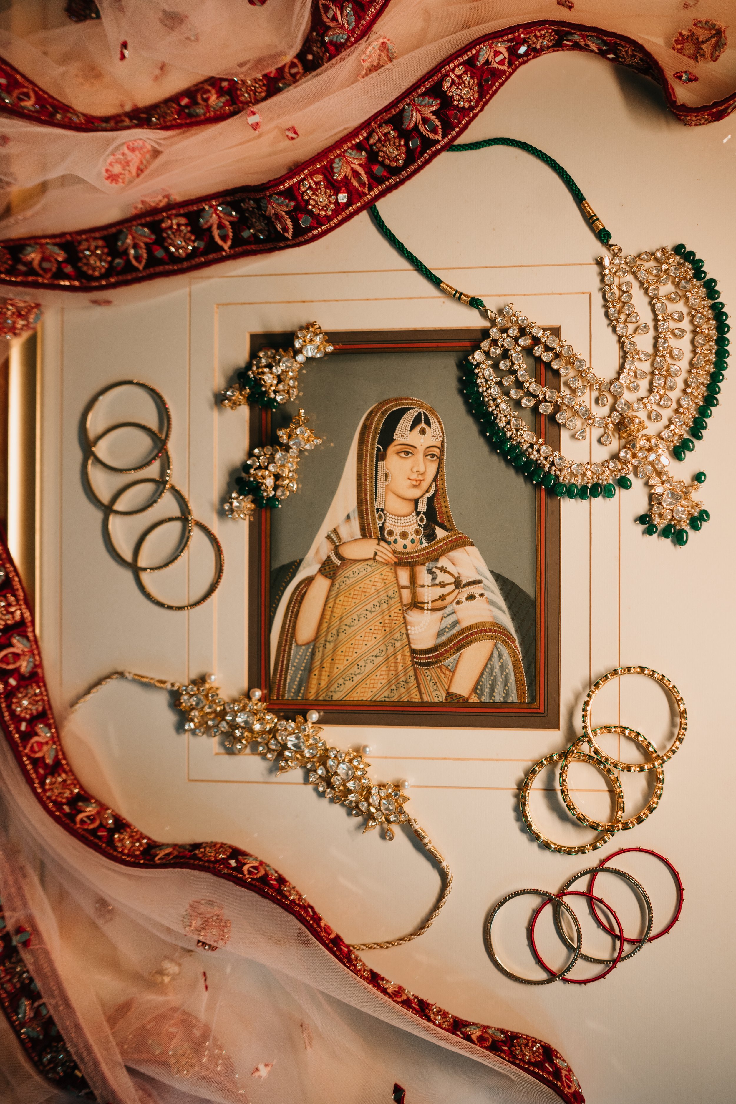 A framed portrait of a woman dressed in traditional Indian attire, surrounded by jewelry including necklaces and bangles, with a colorful embroidered fabric and sheer cloth draped around