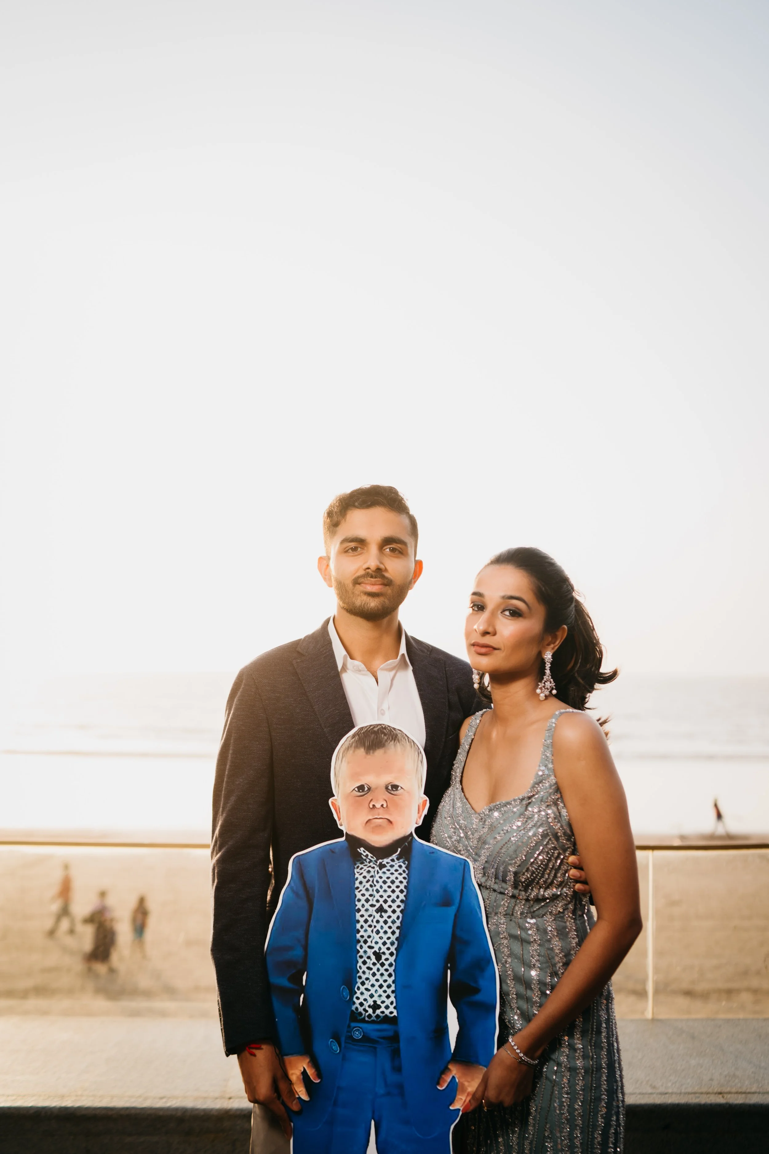 A family of three posing outdoors near the beach during sunset, with the father, mother, and a life-sized cutout of a young boy in a blue suit in front of them.
