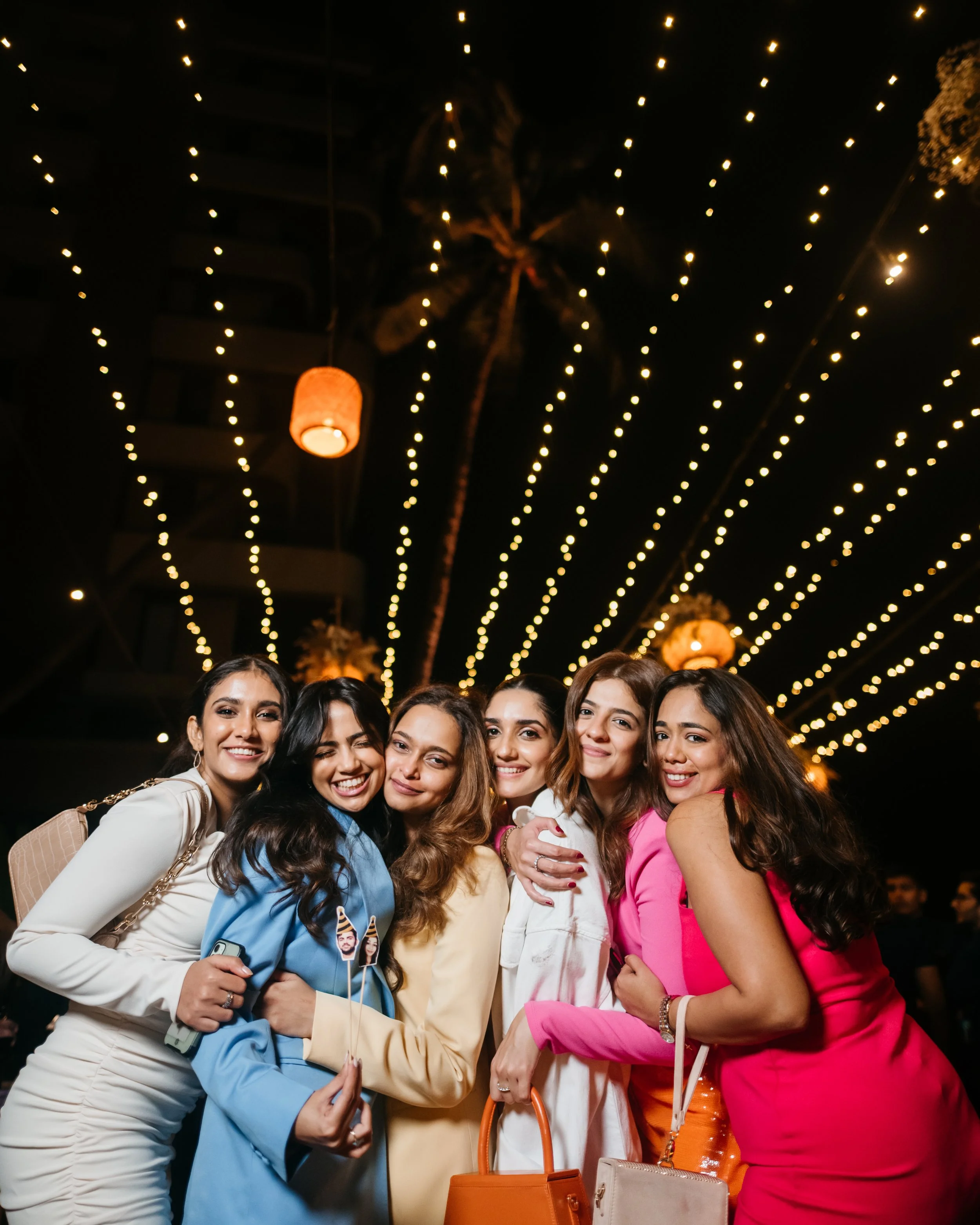 Group of six women smiling and hugging at a nighttime outdoor gathering with string lights and lanterns overhead.