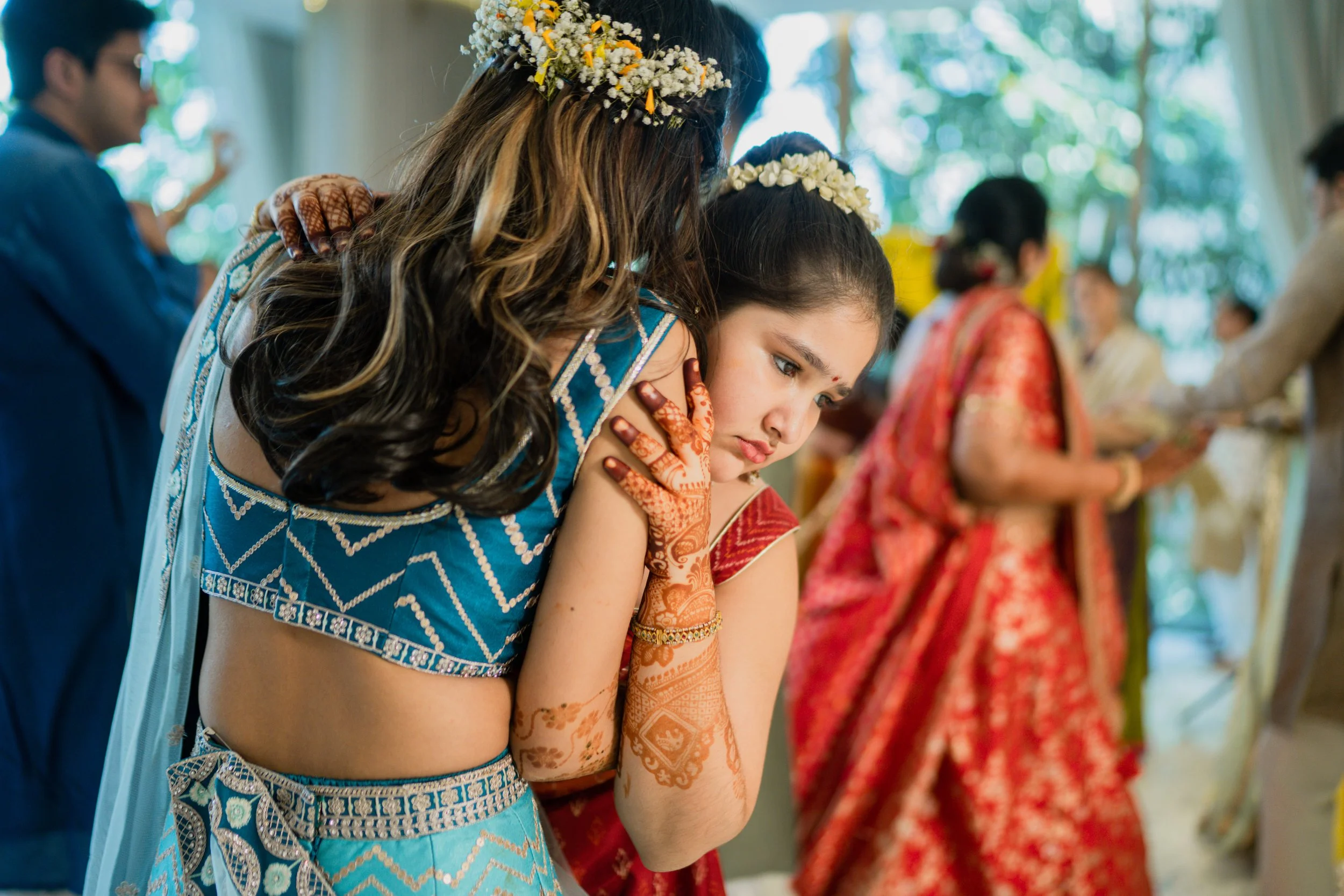 Two women bow their heads in prayer during a traditional Indian wedding ceremony, dressed in colorful sarees and adorned with flower garlands and henna on their hands, with other people participating in the background.