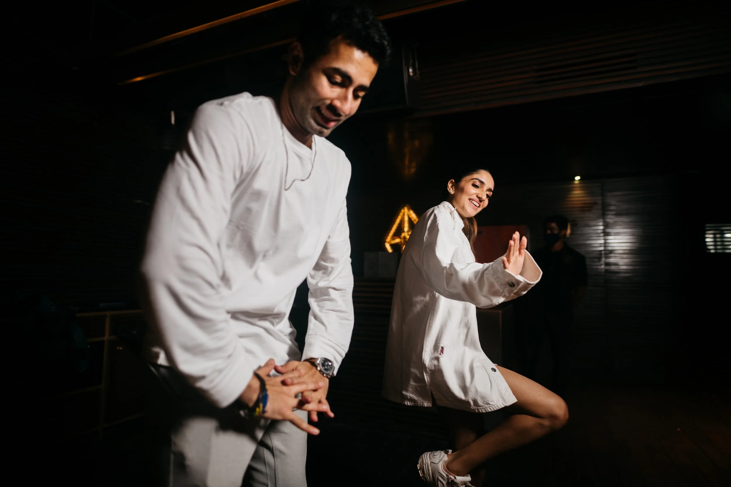 Two people dancing happily in a dark room with wooden walls, both smiling, one man and one woman in casual white clothing.