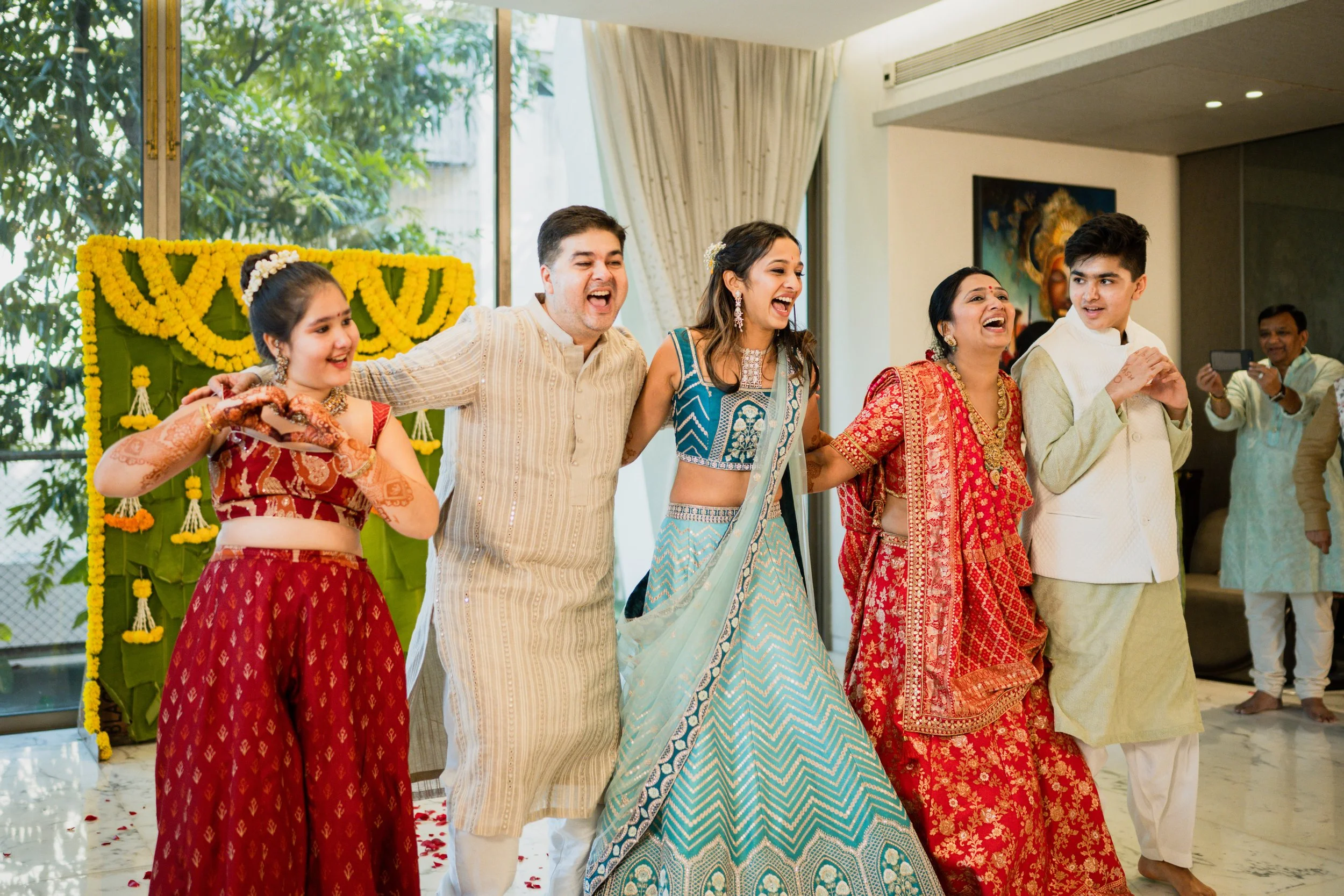 Group of people celebrating, dressed in traditional Indian clothing, smiling and enjoying a festive moment indoors with a decorated background.
