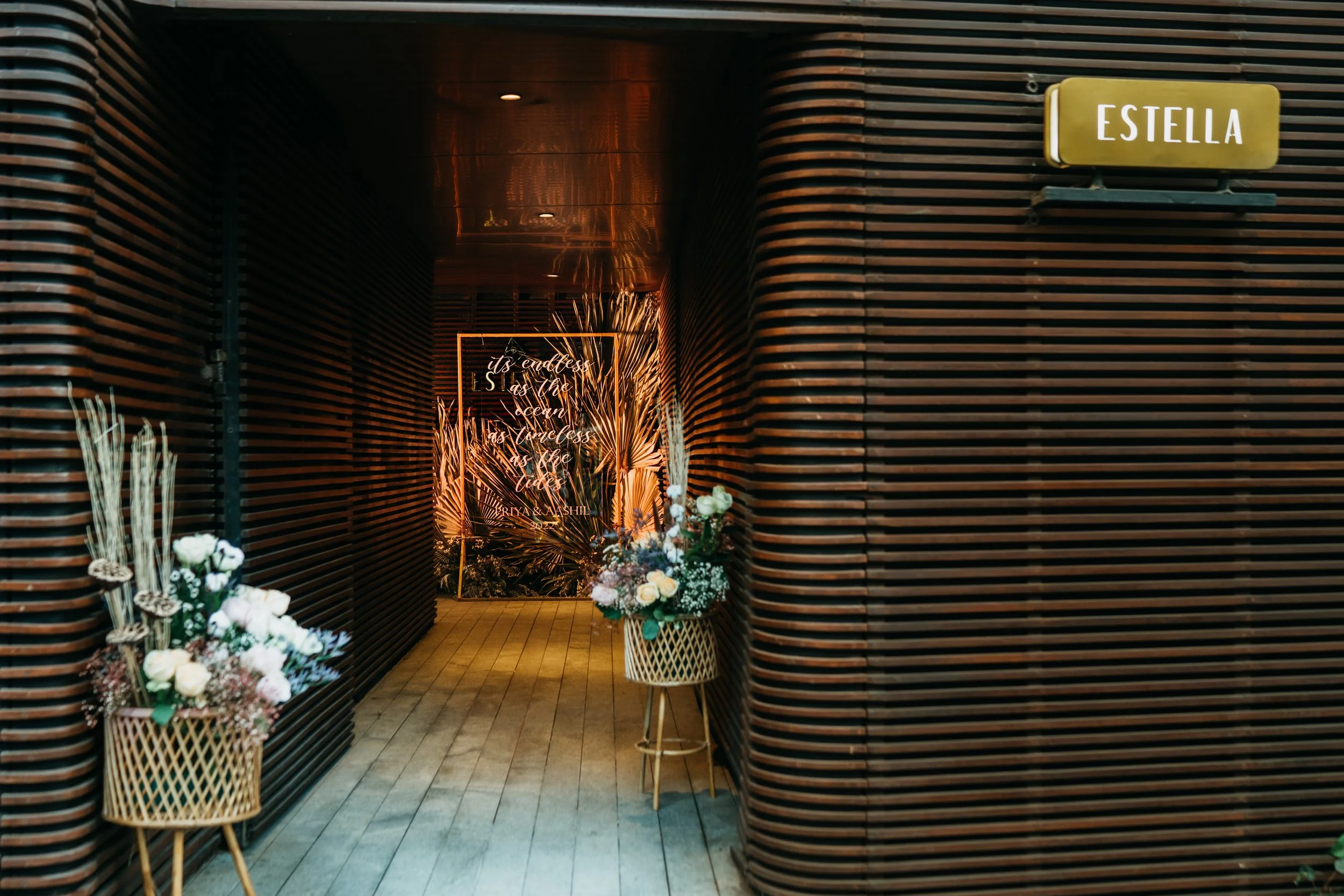 Interior space with wooden slat walls, floral arrangements in woven baskets, illuminated sign with neon script in the background, and a yellow sign labeled 'Estella' on the right.