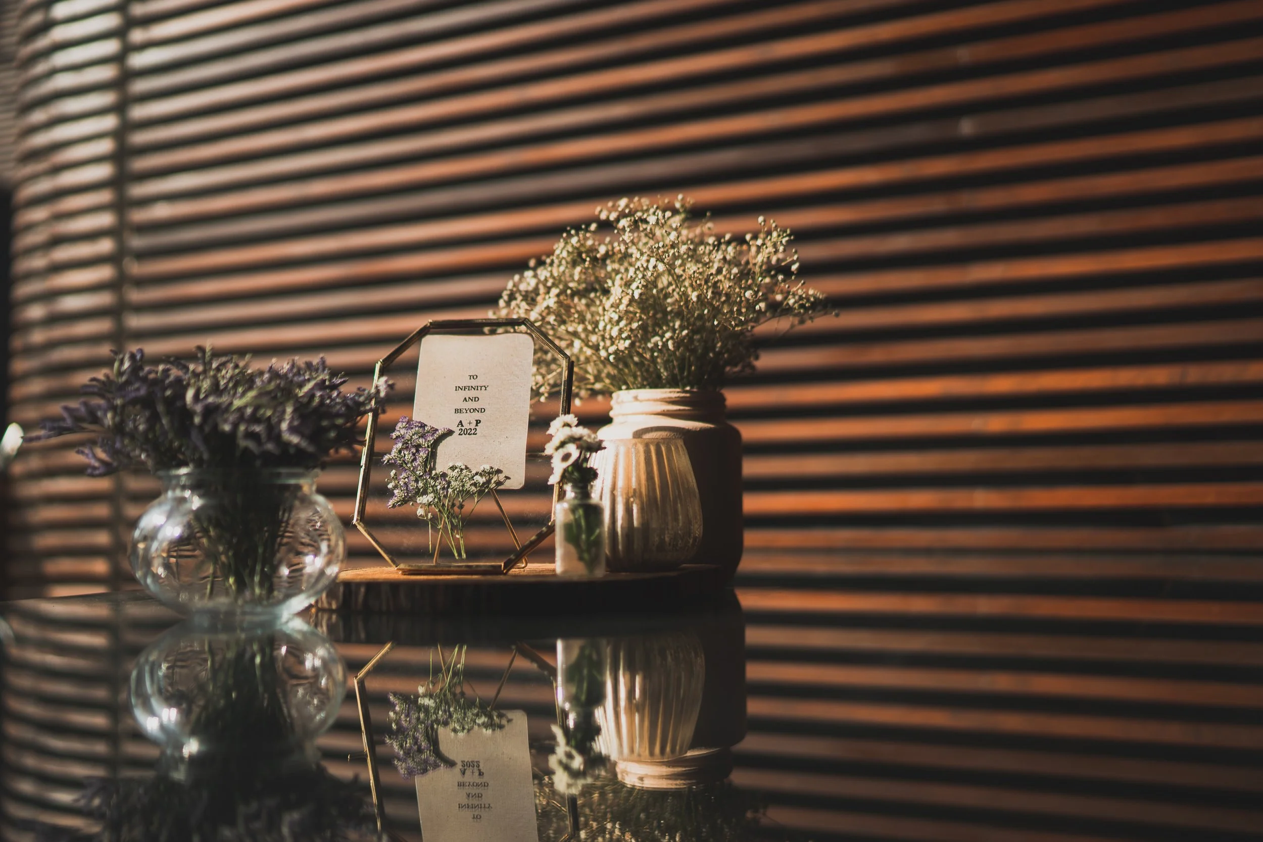 A glass vase and two ceramic vases filled with flowers placed on a reflective surface against a wooden slatted wall. There is also a picture frame with text and small flowers.