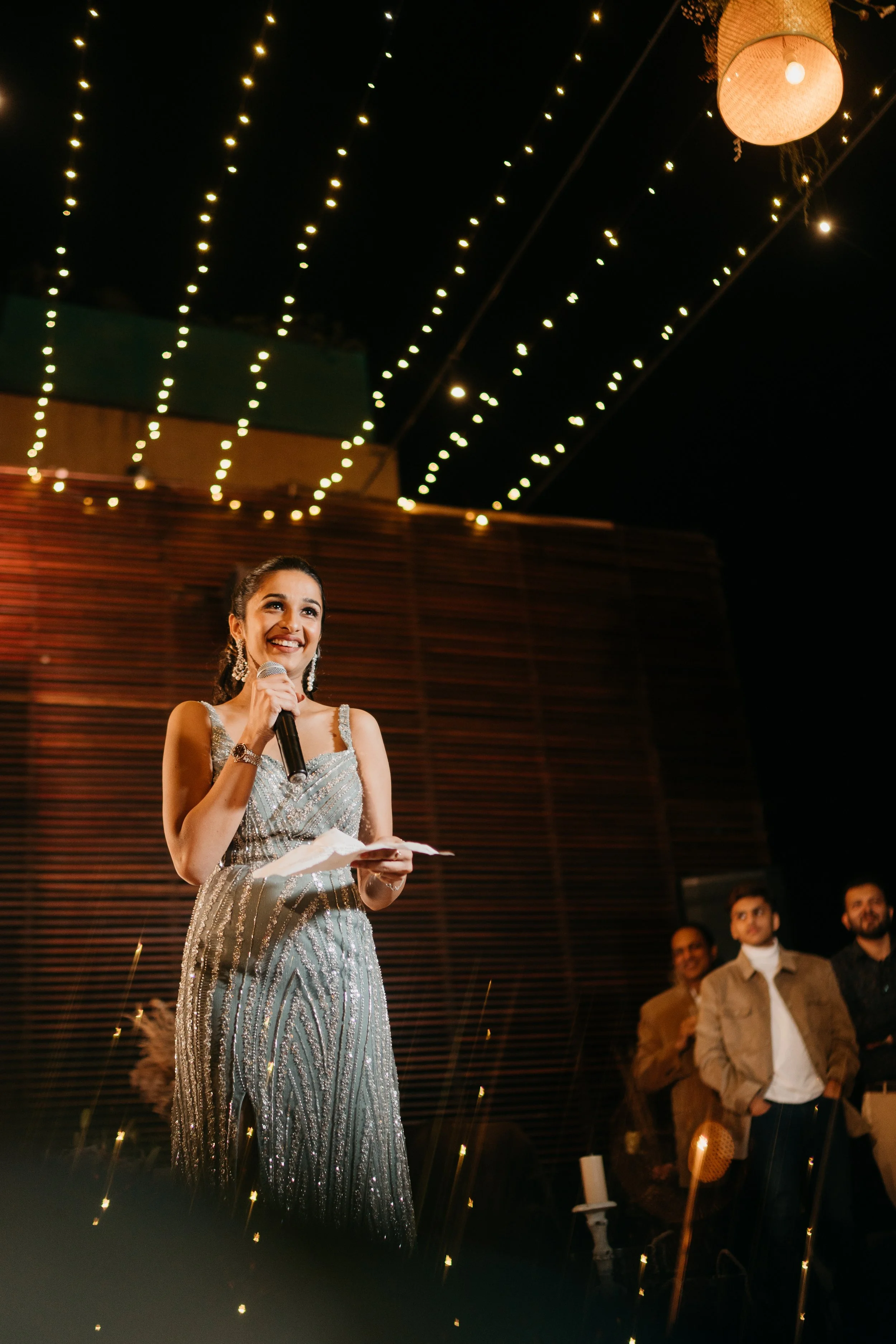 A woman in a sparkly dress holding a microphone and speaking at a celebration or event. Some people are in the background, and string lights decorate the venue ceiling at night.