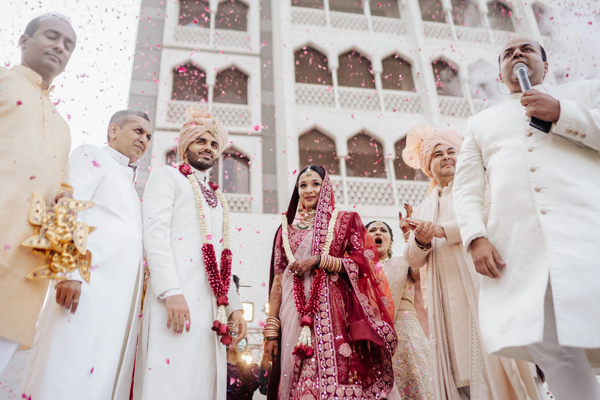 Indian wedding celebration with a bride in a red traditional sari and groom in white, surrounded by family members, as flower petals fall.