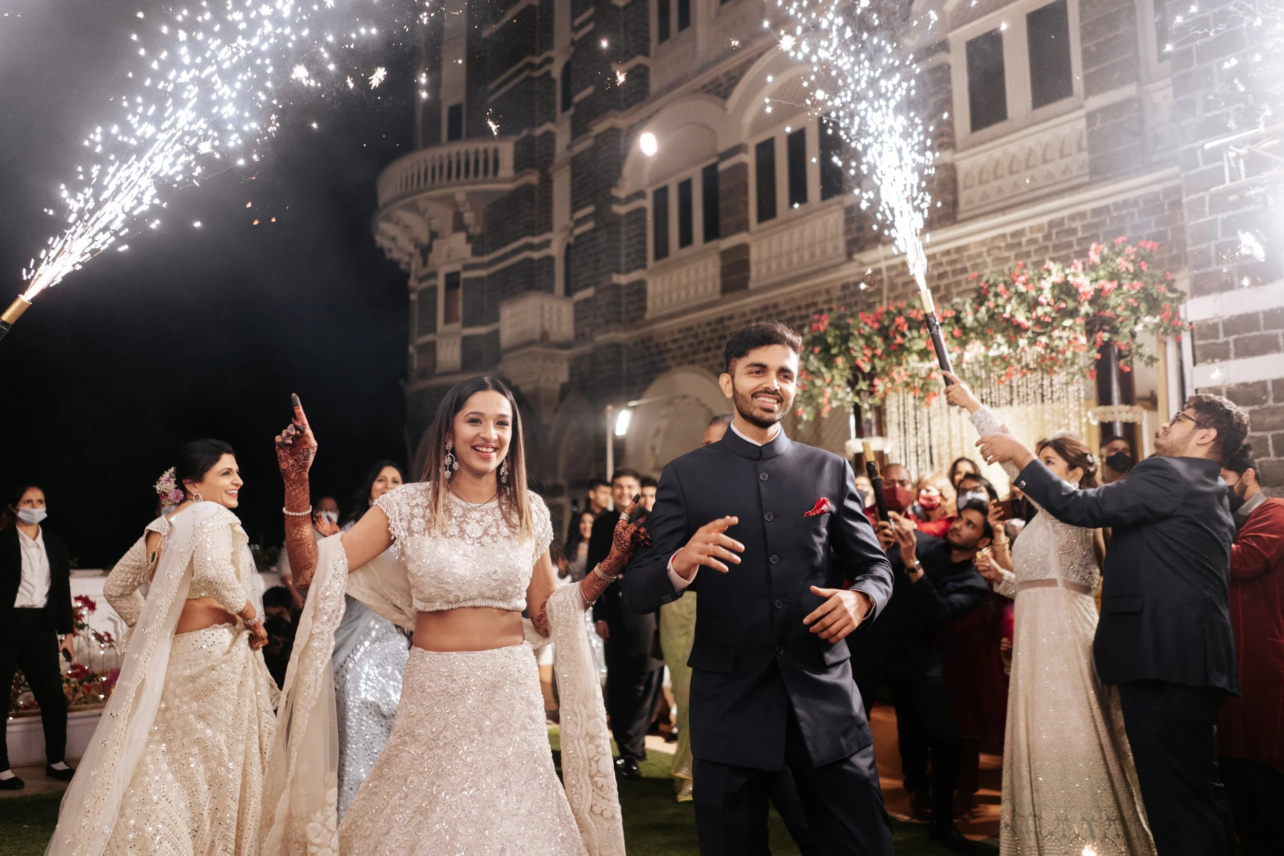 A wedding celebration at night with a bride in traditional attire, a groom in a black suit, and guests holding sparklers in front of a large, ornate building.