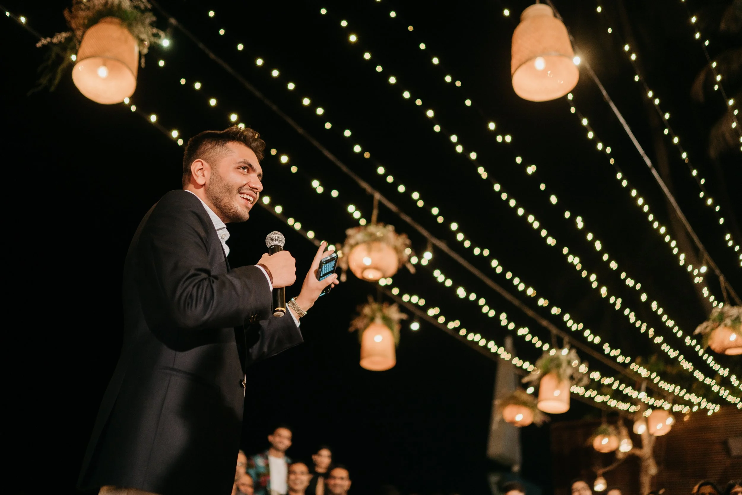 A man in a black suit holding a microphone and a smartphone, speaking to an outdoor gathering at night with hanging lanterns and string lights overhead, and a group of people in the background.