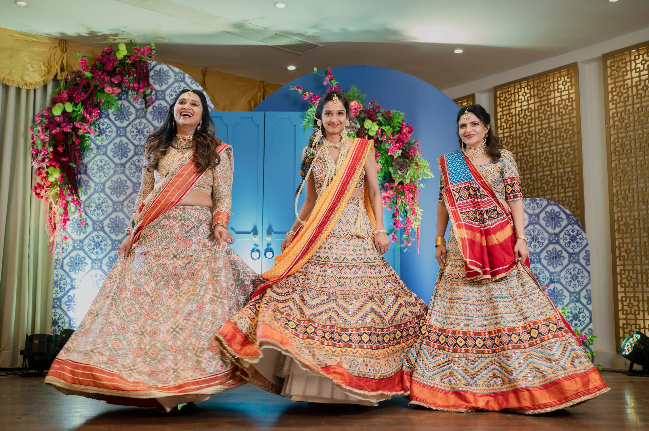 Three women in colorful traditional Indian attire dancing on stage with floral decorations and a blue backdrop.