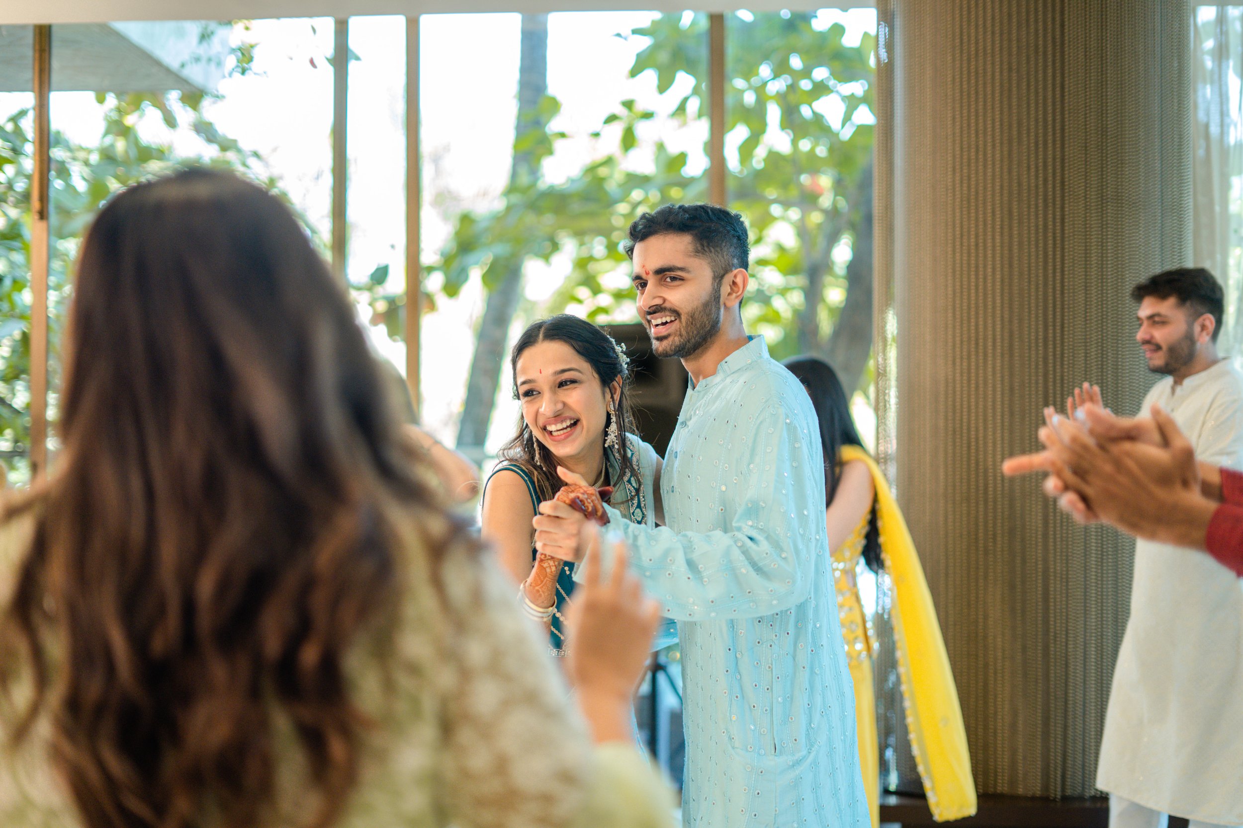 A group of people celebrating at a festive event, with a young man and woman dancing and smiling, surrounded by others clapping and enjoying the occasion, in a room with large windows and greenery outside.