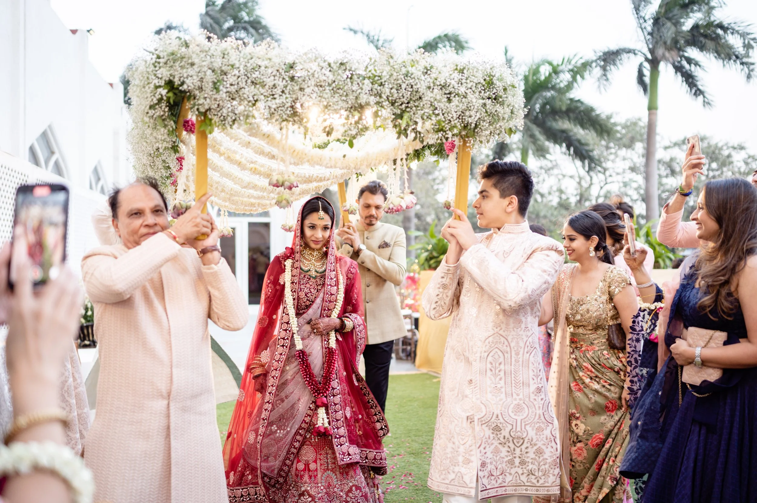 Indian bride in red traditional wedding attire under a floral canopy, surrounded by family and friends celebrating outdoors.
