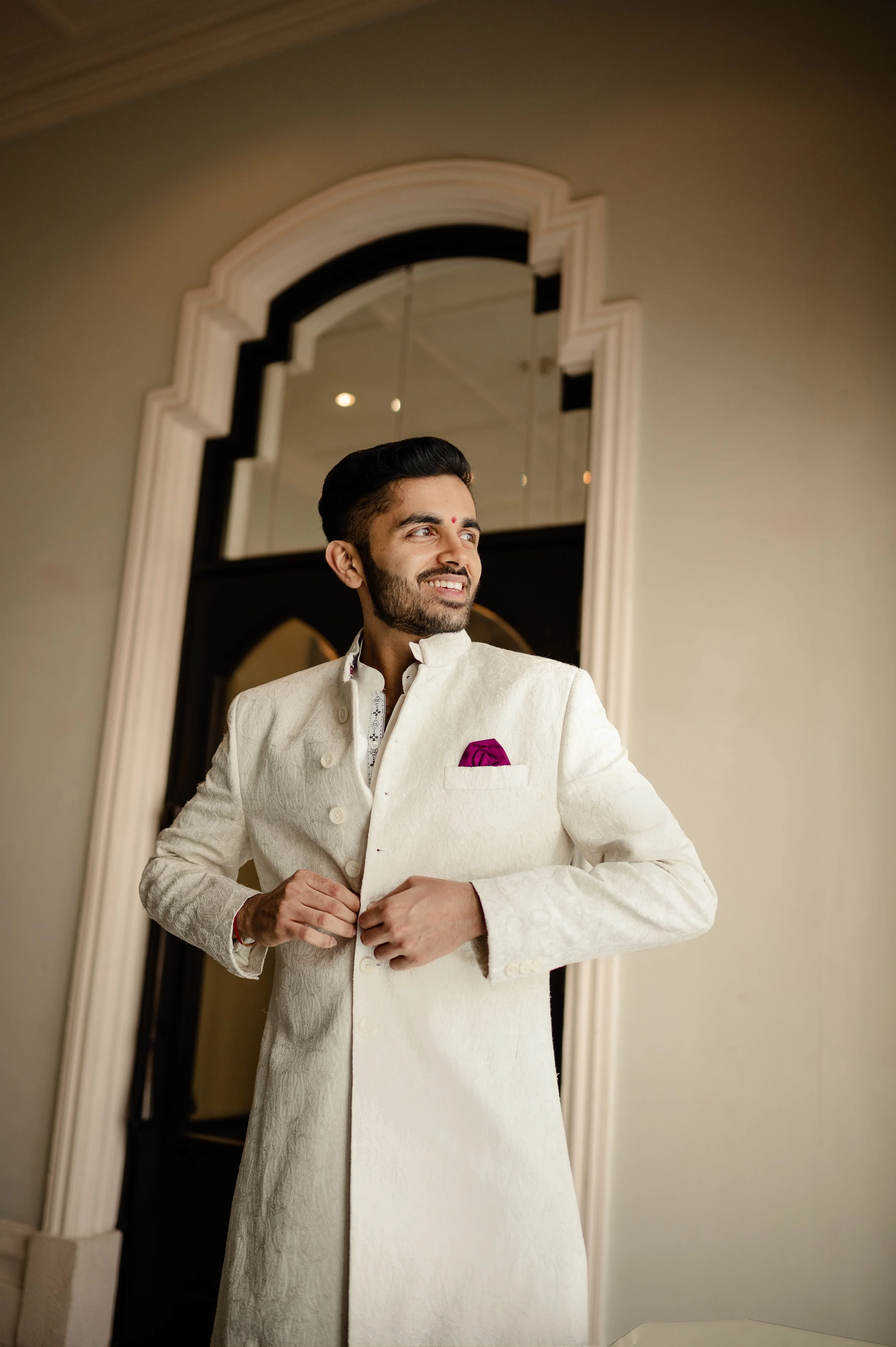 A man dressed in a white traditional Indian outfit, adjusting his buttons, standing in front of a decorative mirror, with a bright smile on his face.