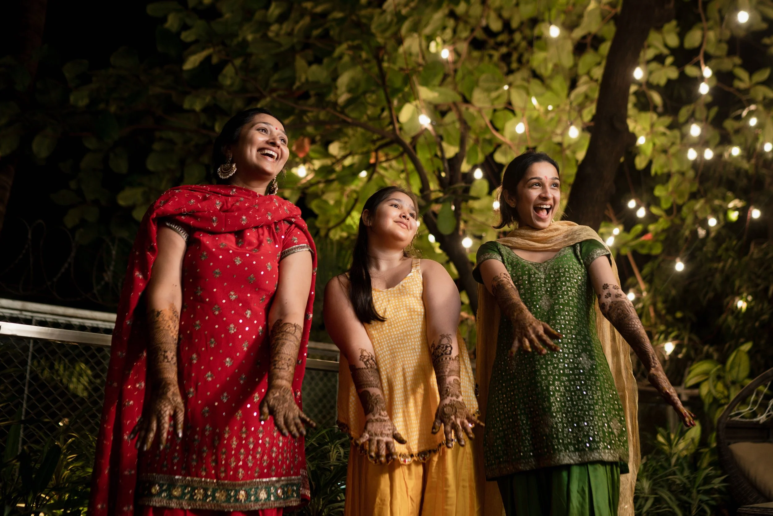 Three women and a girl in traditional Indian attire with henna on their hands, smiling and enjoying a night outdoors under string lights.