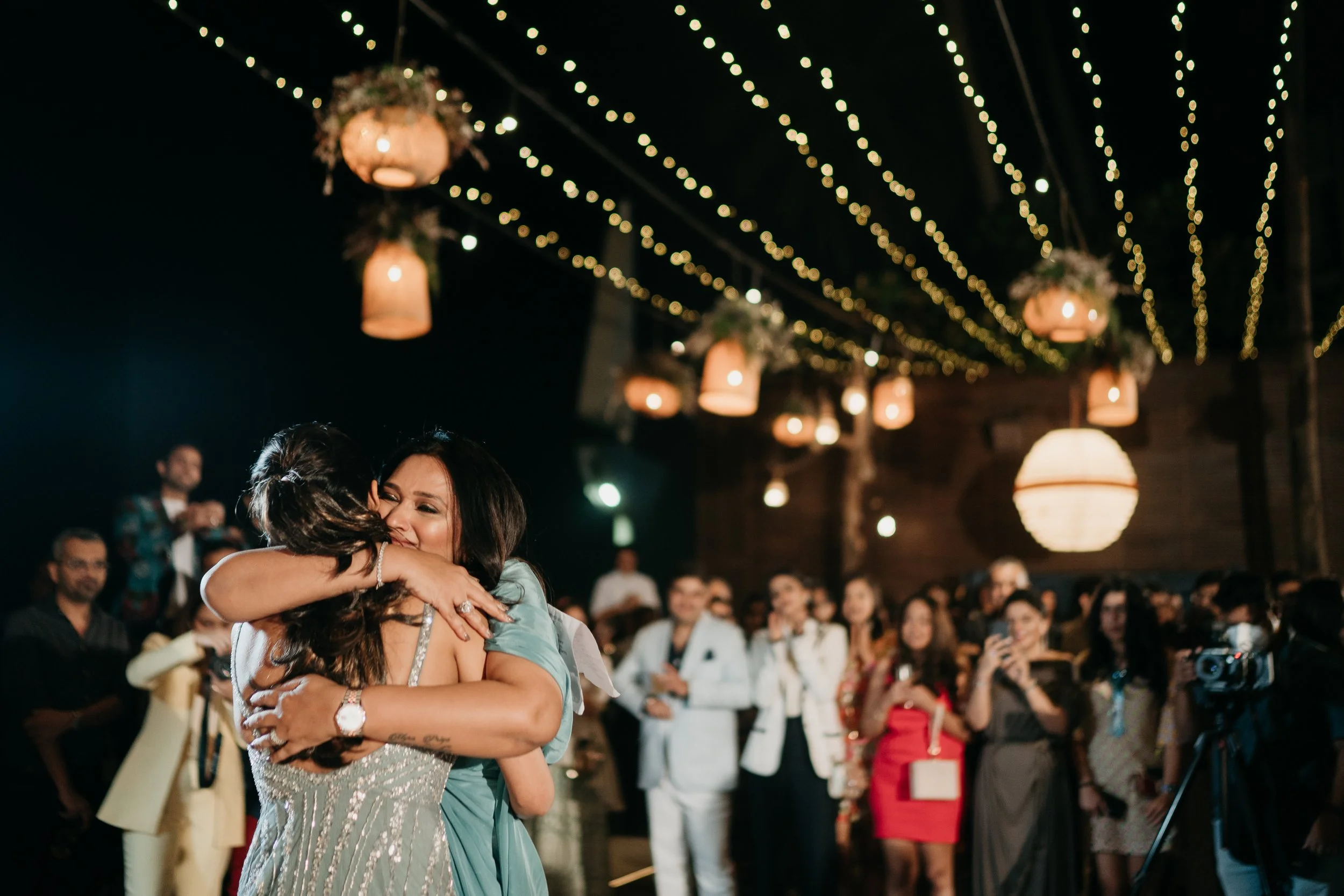 Two women hugging and smiling at a celebration in a decorated indoor venue with hanging lights and an audience in the background.