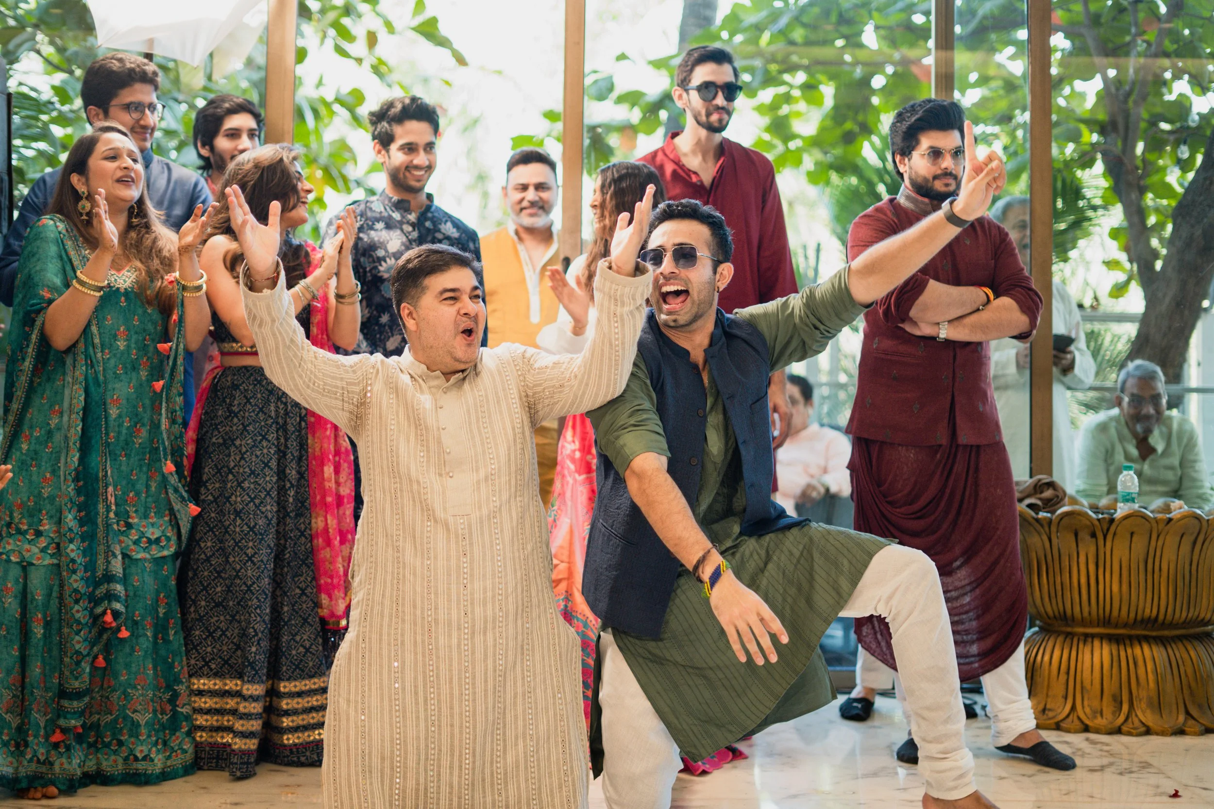 A group of people celebrating and dancing at a festive gathering, with some clapping and others singing or enjoying the moment, indoors with large glass windows and green foliage outside.