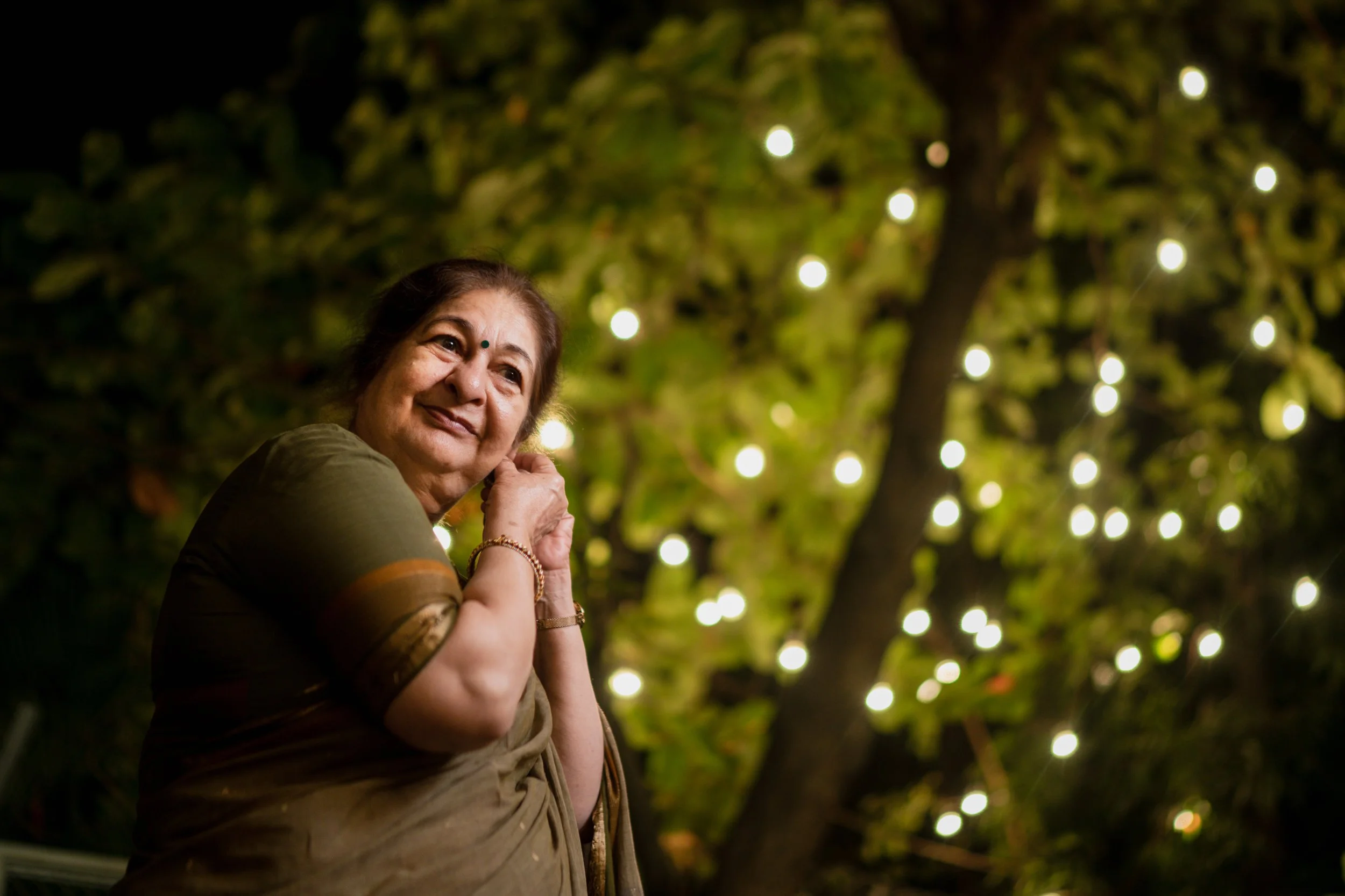 A woman in traditional Indian attire enjoying outdoors at night, illuminated by string lights in the background.