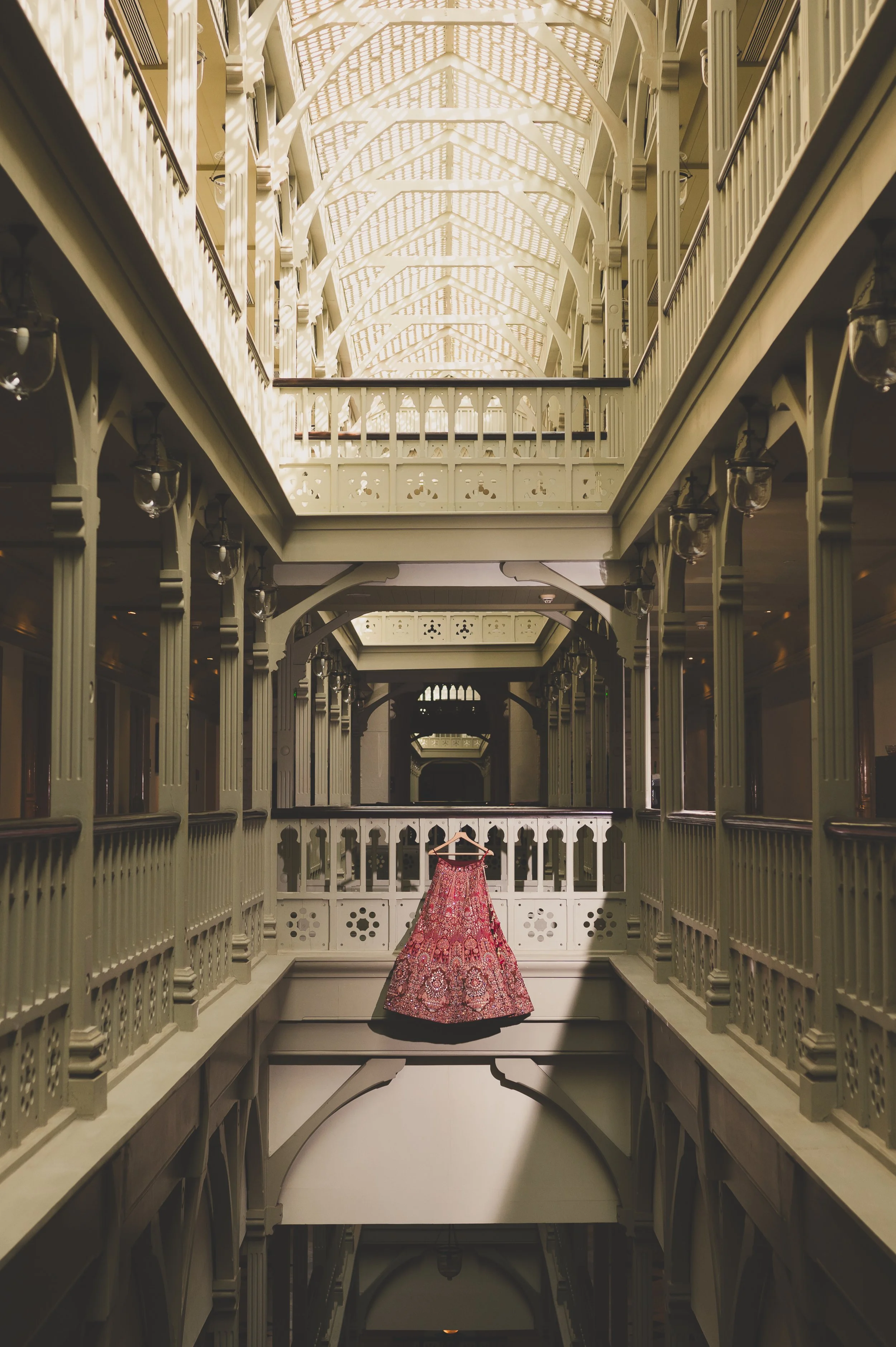 A pink floral dress hanging on a hanger in the center of an ornate, multi-story interior with cream-colored railings and decorative woodwork.