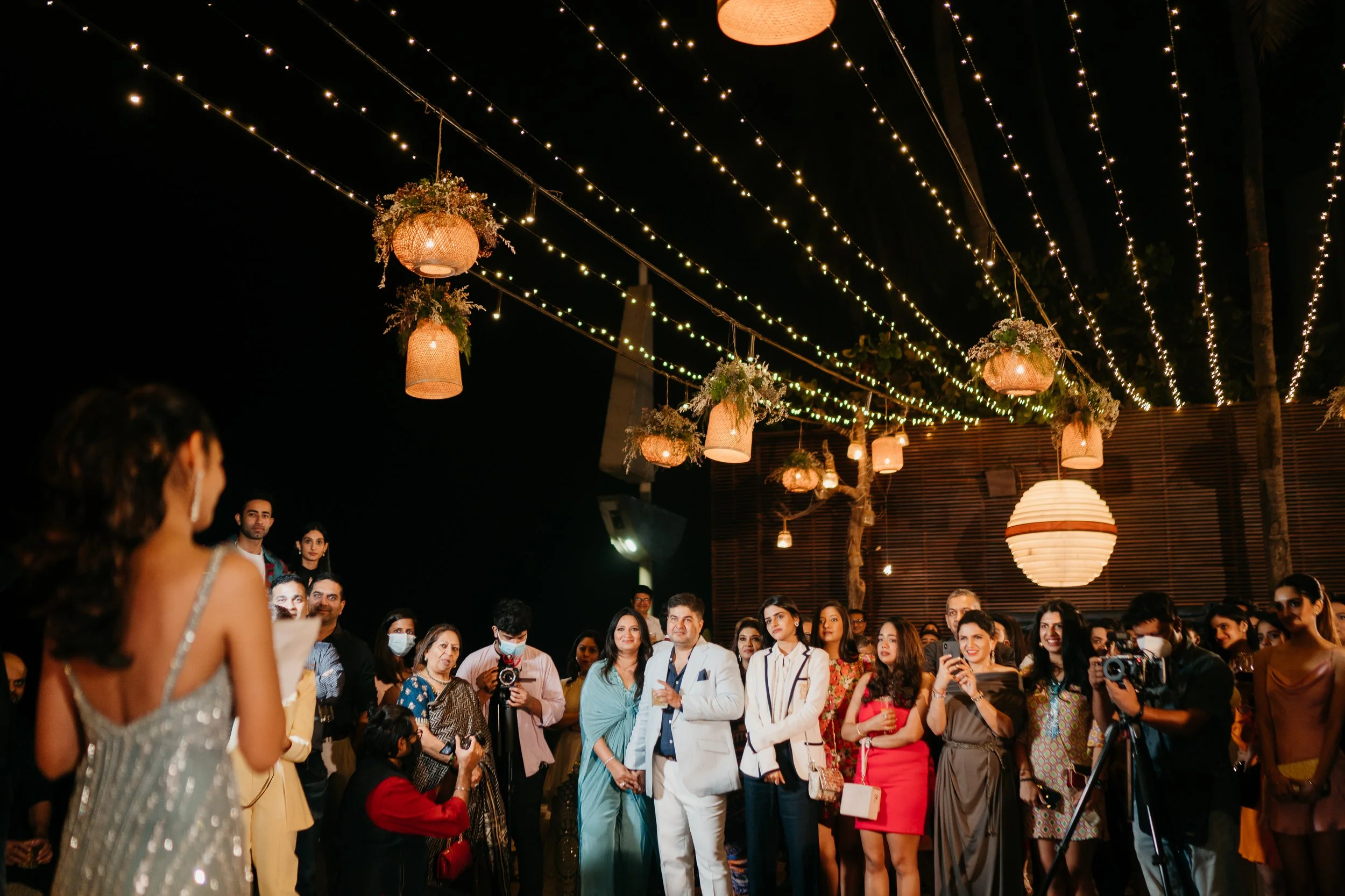 Guests attending an outdoor nighttime event decorated with hanging lanterns and string lights.