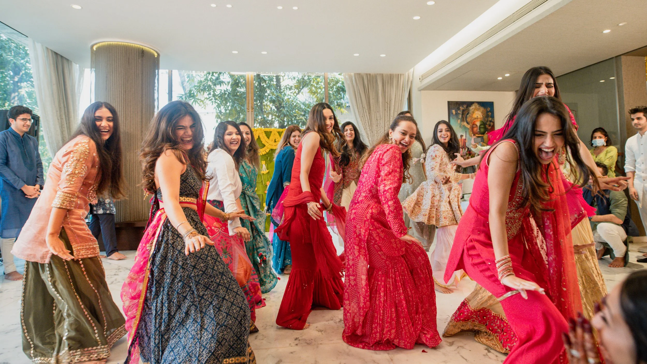 A group of women dressed in colorful traditional Indian attire dancing and celebrating inside a decorated room.
