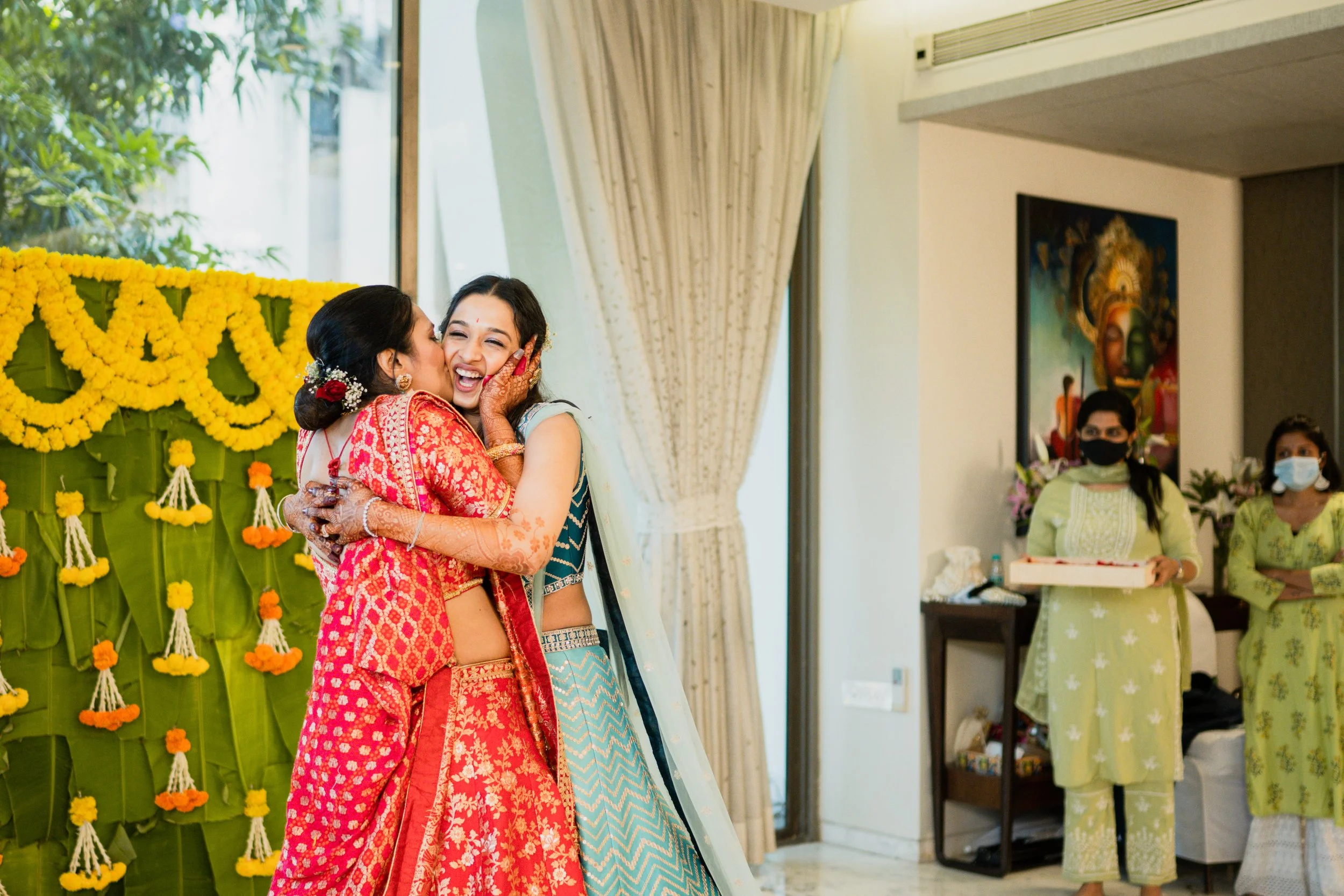 Women celebrating at a traditional Indian wedding, with one woman in a red saree hugging another in a blue-green saree, both smiling and joyful, surrounded by decorated floral backdrop and guests wearing masks in indoor setting.