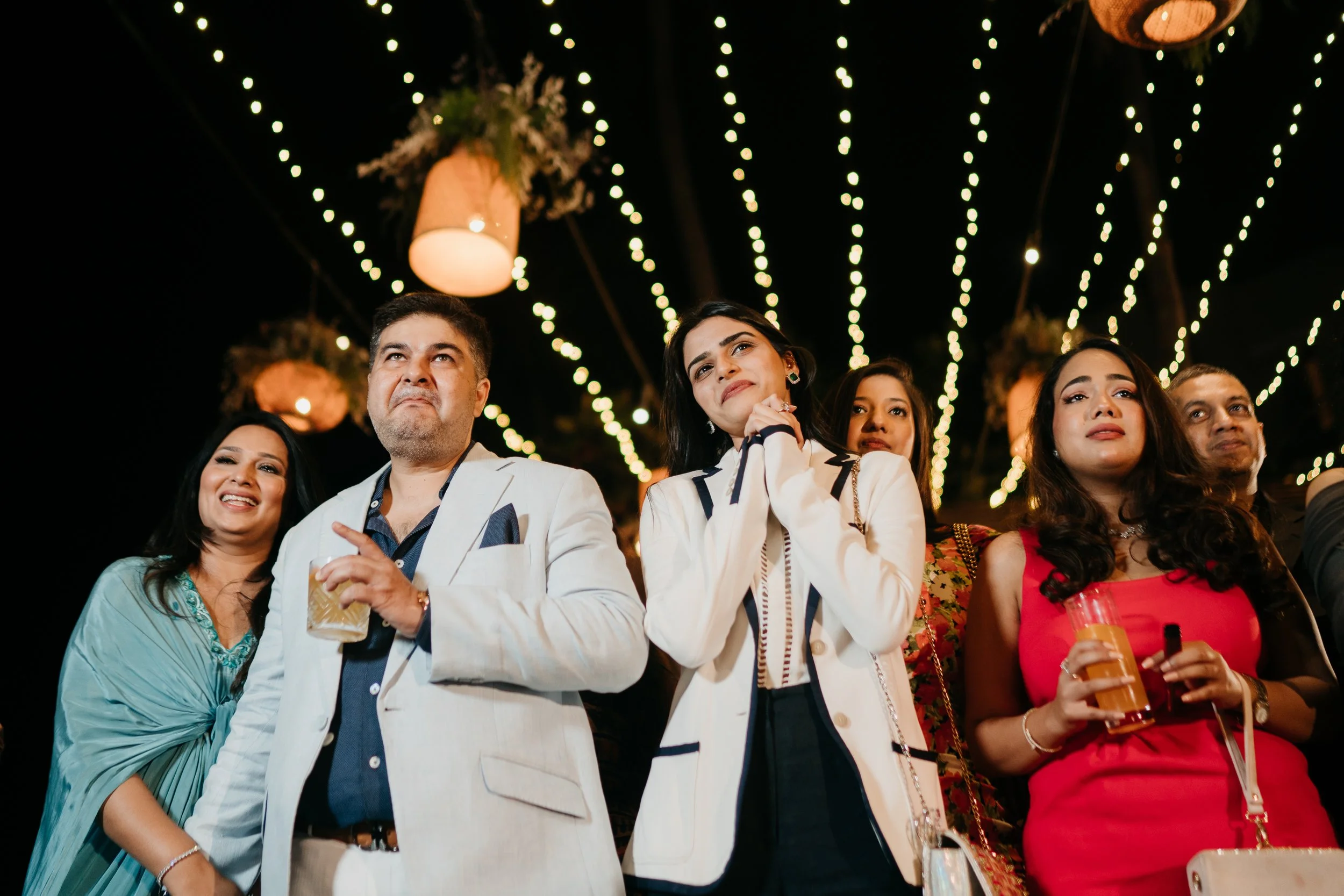 Group of six people at a celebration, standing outdoors at night under string lights and hanging lanterns, some smiling, some looking serious, holding drinks.