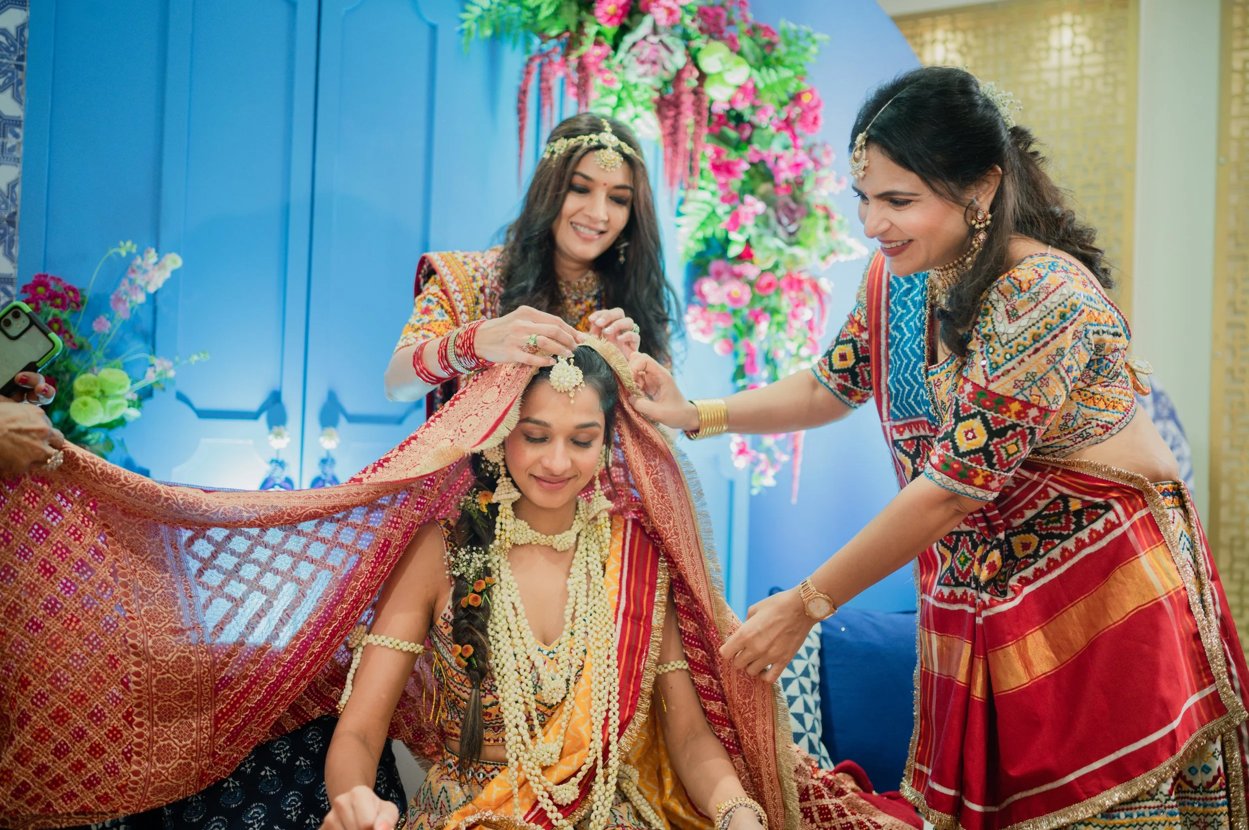 A traditional Indian wedding ceremony with the bride sitting with closed eyes, surrounded by women who are adjusting her jewelry and veil, decorated with colorful flowers and vibrant attire.