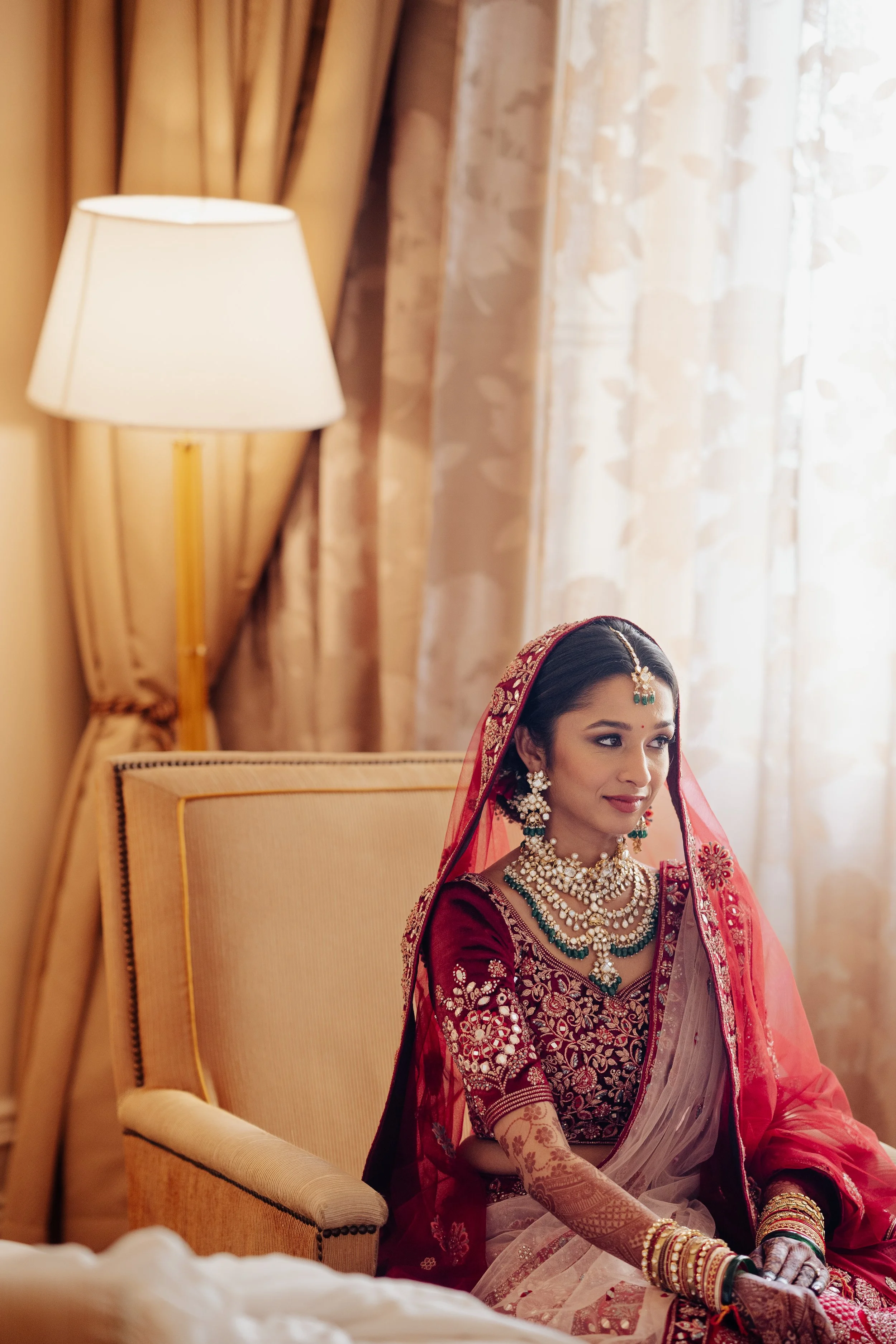 A woman dressed in traditional South Asian bridal attire sitting on a beige armchair in a warmly lit room with curtains and a lamp in the background.