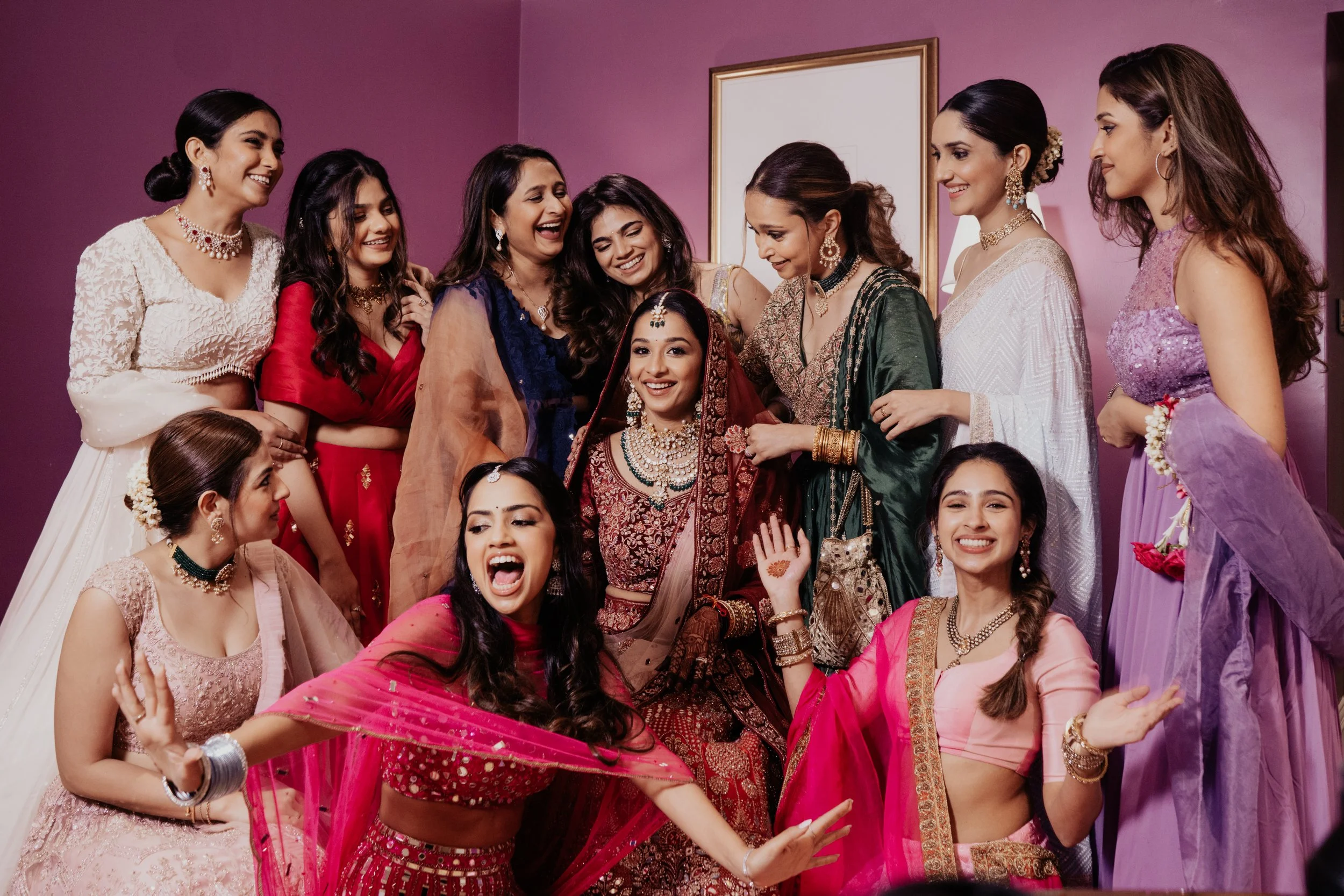 A group of women dressed in colorful traditional Indian attire, celebrating and smiling at a wedding ceremony.