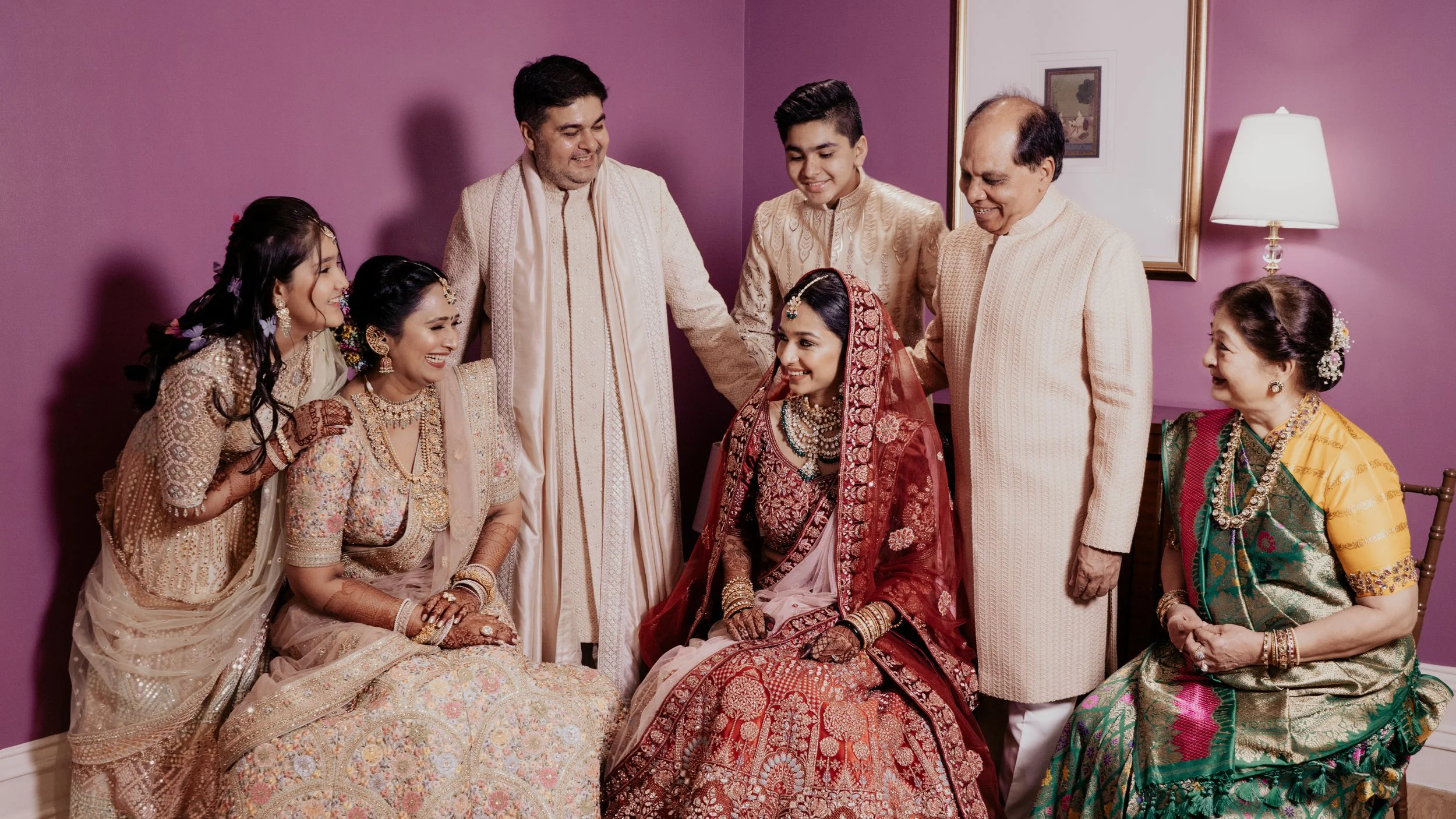 A group of people dressed in traditional Indian attire gathered around a seated woman in bridal wedding jewelry, smiling and celebrating together in a warmly lit room with a purple wall and a framed picture.