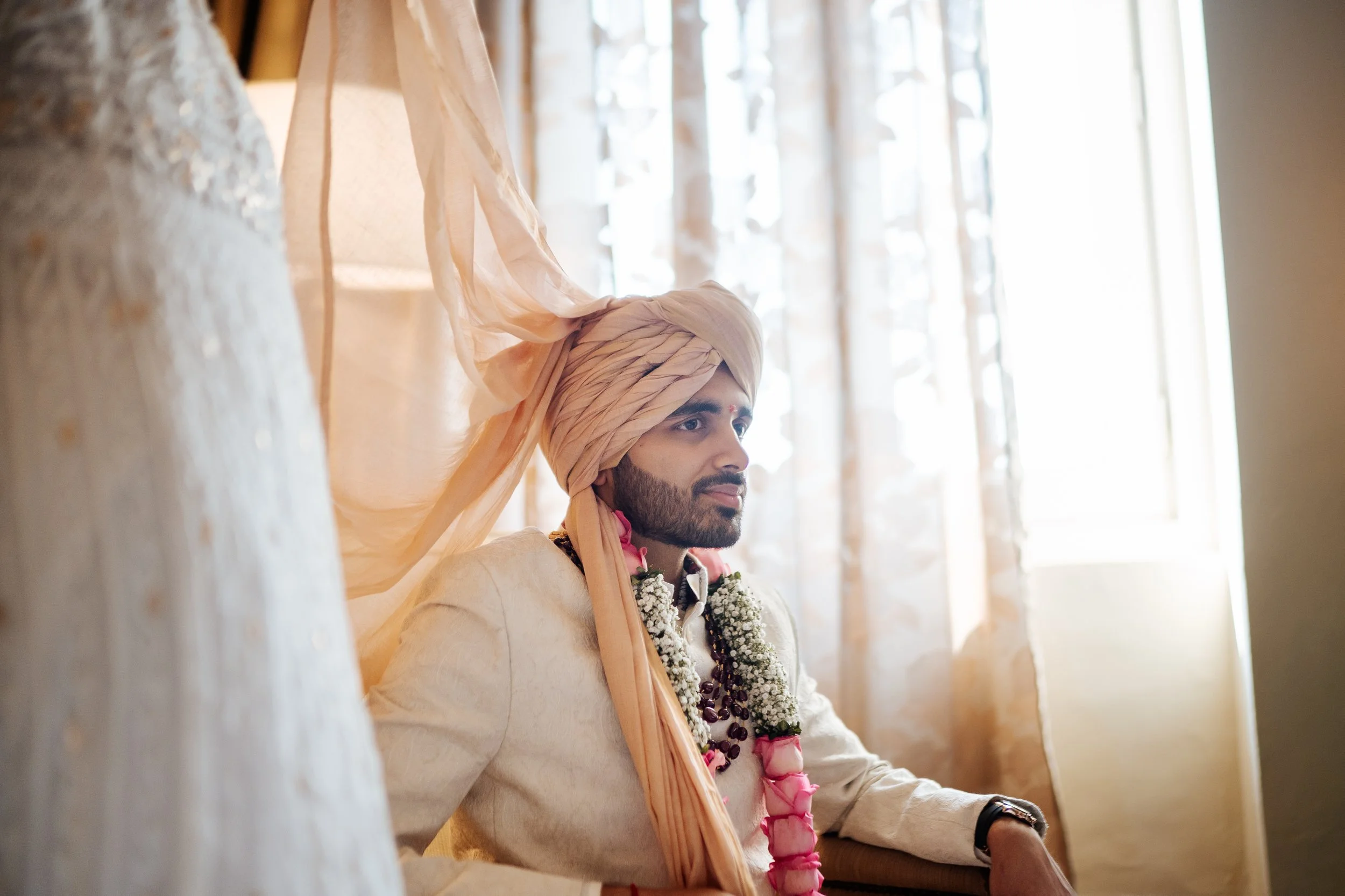 Indian groom wearing traditional attire and a turban, sitting indoors near a window with curtains.