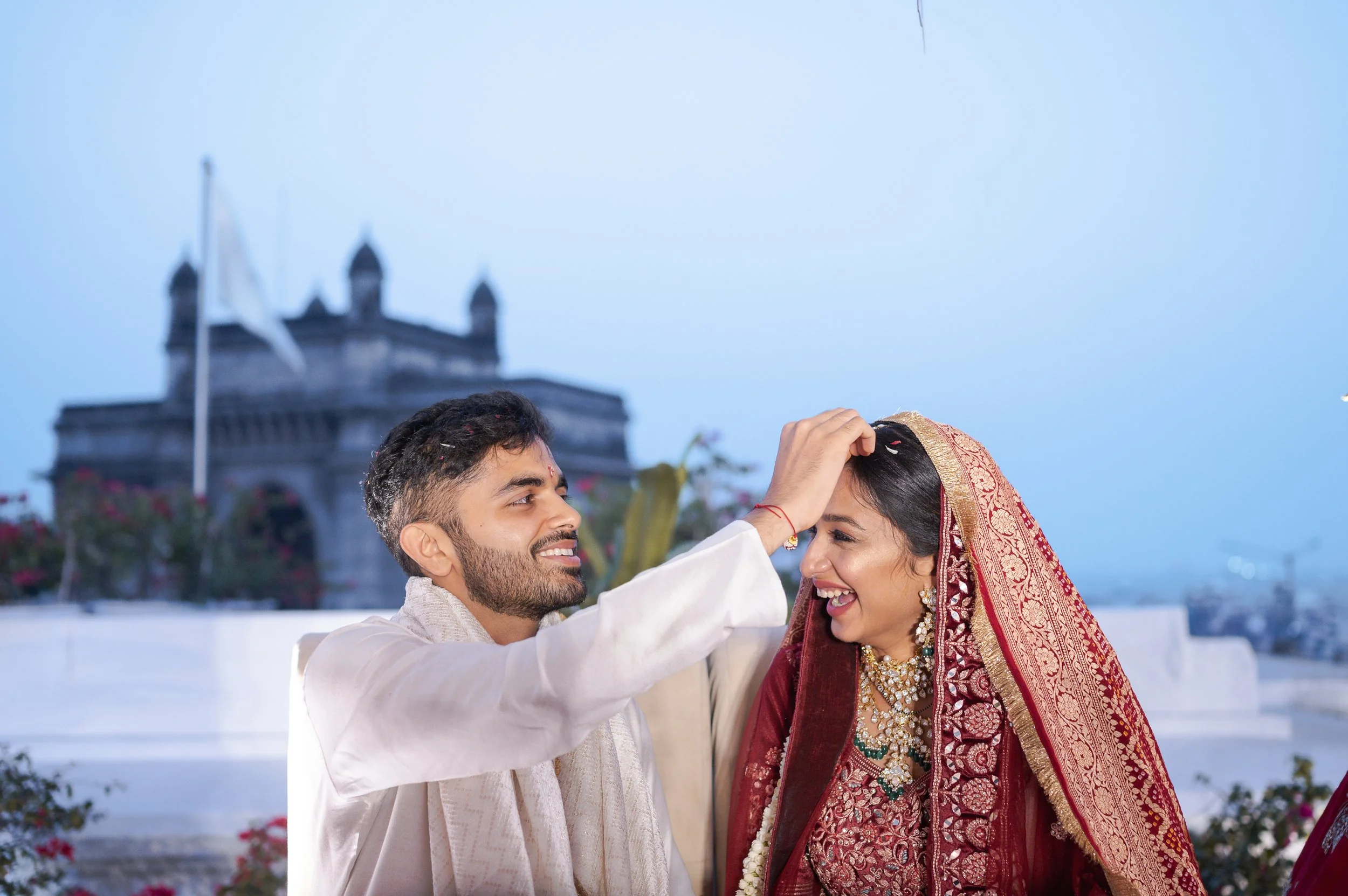 A couple dressed in traditional Indian wedding attire, smiling and celebrating on a terrace with a historical building in the background.