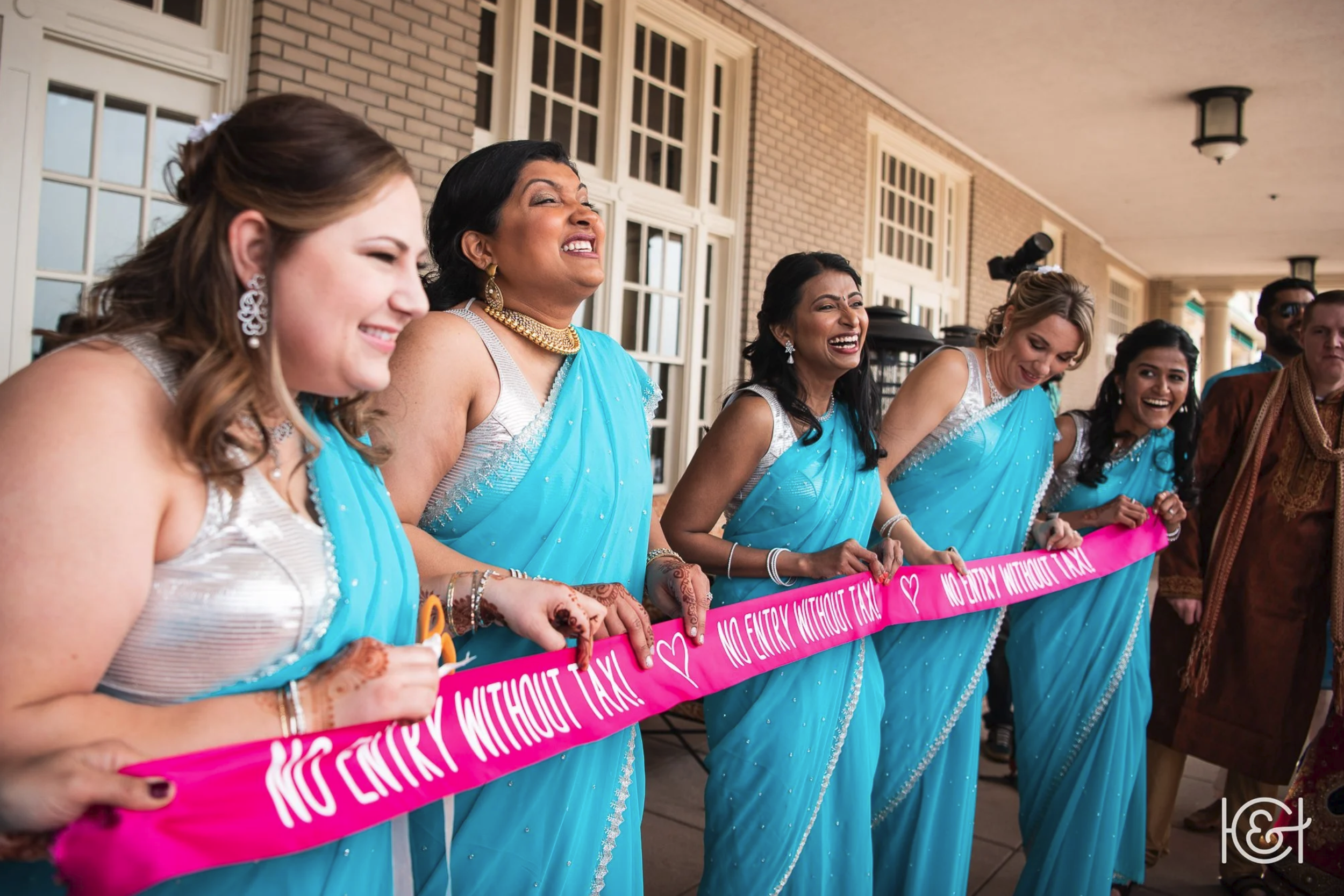 Women dressed in blue sarees participating in a ribbon-cutting ceremony, holding a pink ribbon that reads 'NO ENTRY WITHOUT TAX' on a porch or outdoor veranda.