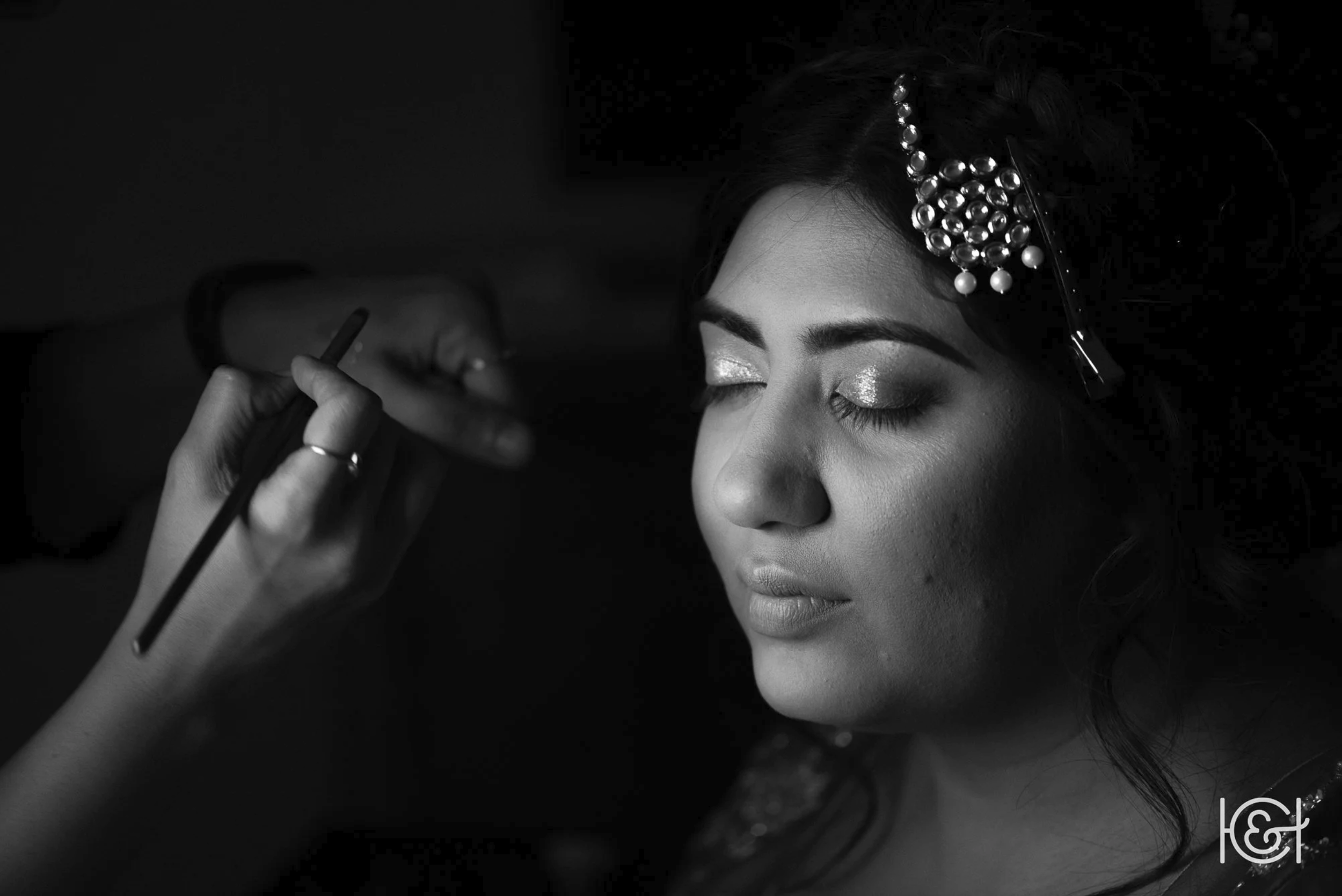A woman with closed eyes having makeup applied by a person holding a brush, with decorative pearl and rhinestone hair accessories, in black and white.
