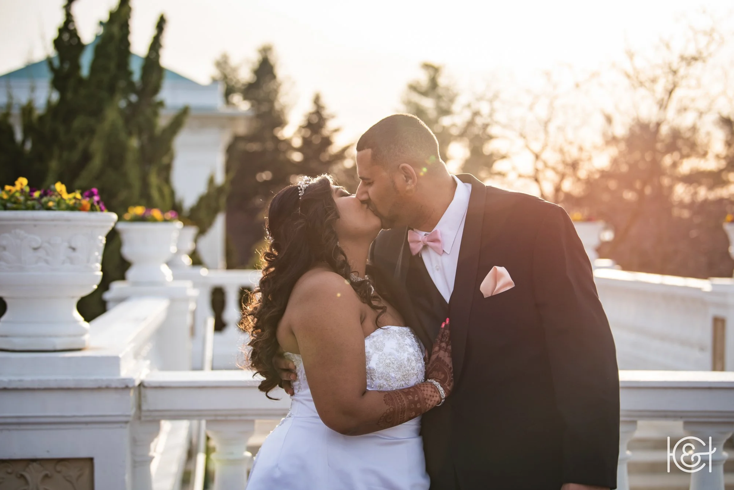 A newlywed couple sharing a kiss outdoors during sunset, with trees and decorative white balustrades in the background.