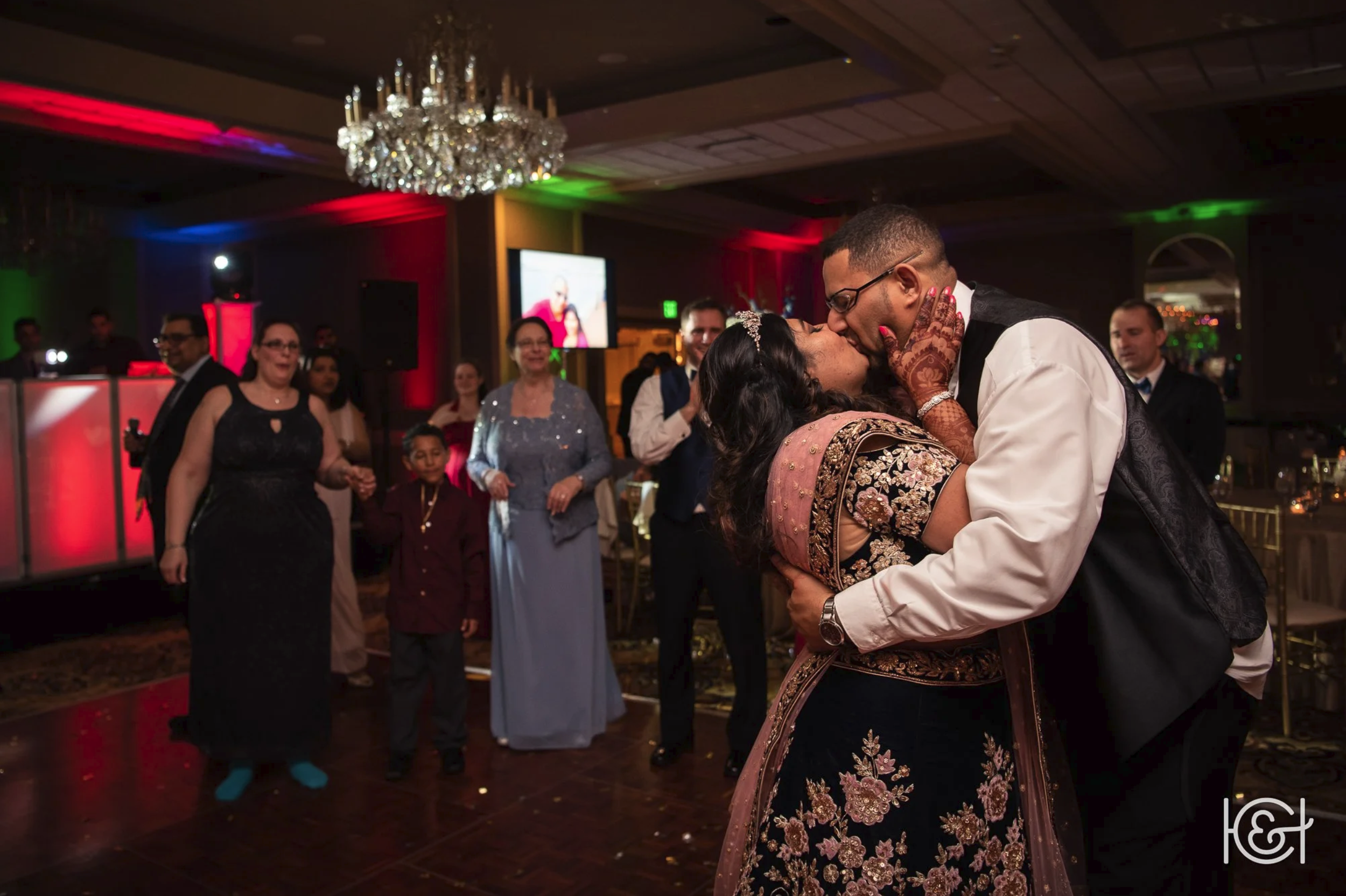 Couple sharing a kiss during a celebration, with guests watching fondly in a decorated banquet hall with a chandelier and colorful lighting.
